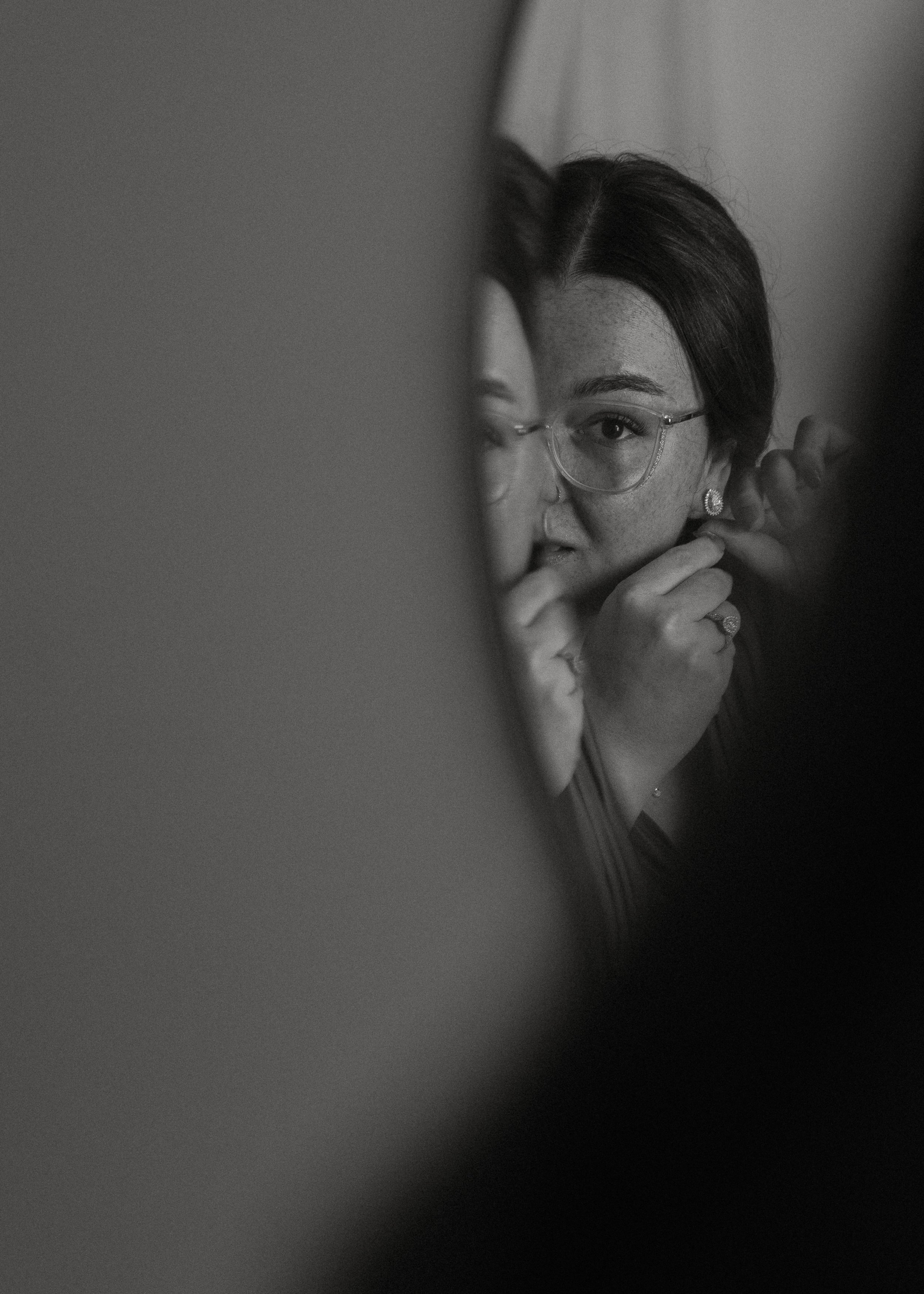A woman wearing glasses and earrings, partially visible through a blurred foreground, adjusting her earring or hair in front of a mirror.