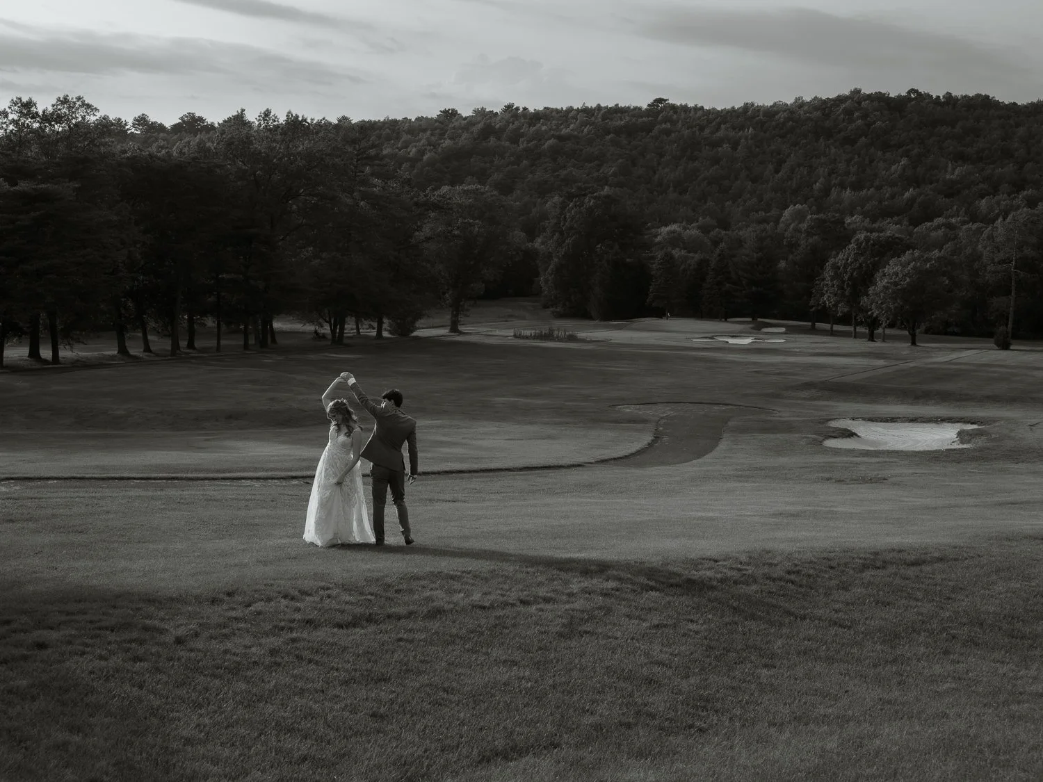 A couple in wedding attire dancing on a golf course with trees and hills in the background, black and white photo.