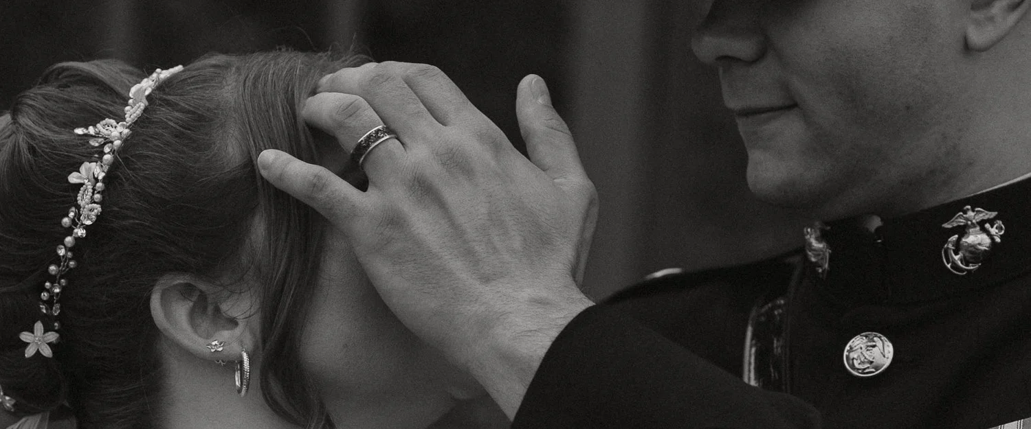 A man places his hand on a young woman's head during a wedding ceremony. The young woman has flowers in her hair, earrings, and a tiara. The scene is tender and intimate.