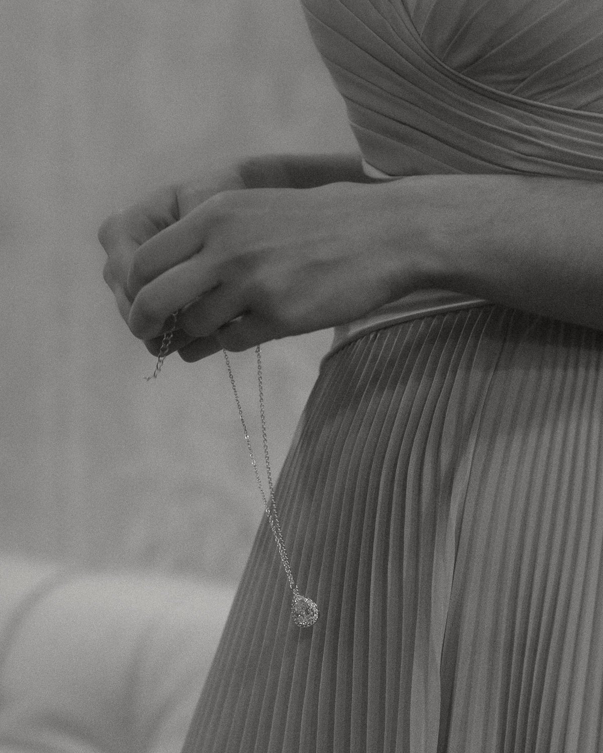 Close-up of a woman holding a delicate necklace with a teardrop-shaped pendant, wearing a pleated, textured dress in black and white.