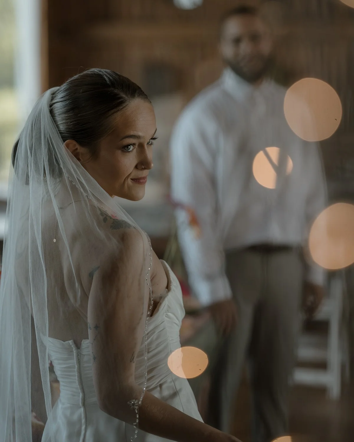 A bride with a veil and tattooed arm looks to the side inside a rustic wooden wedding venue, with a groom standing in the background.