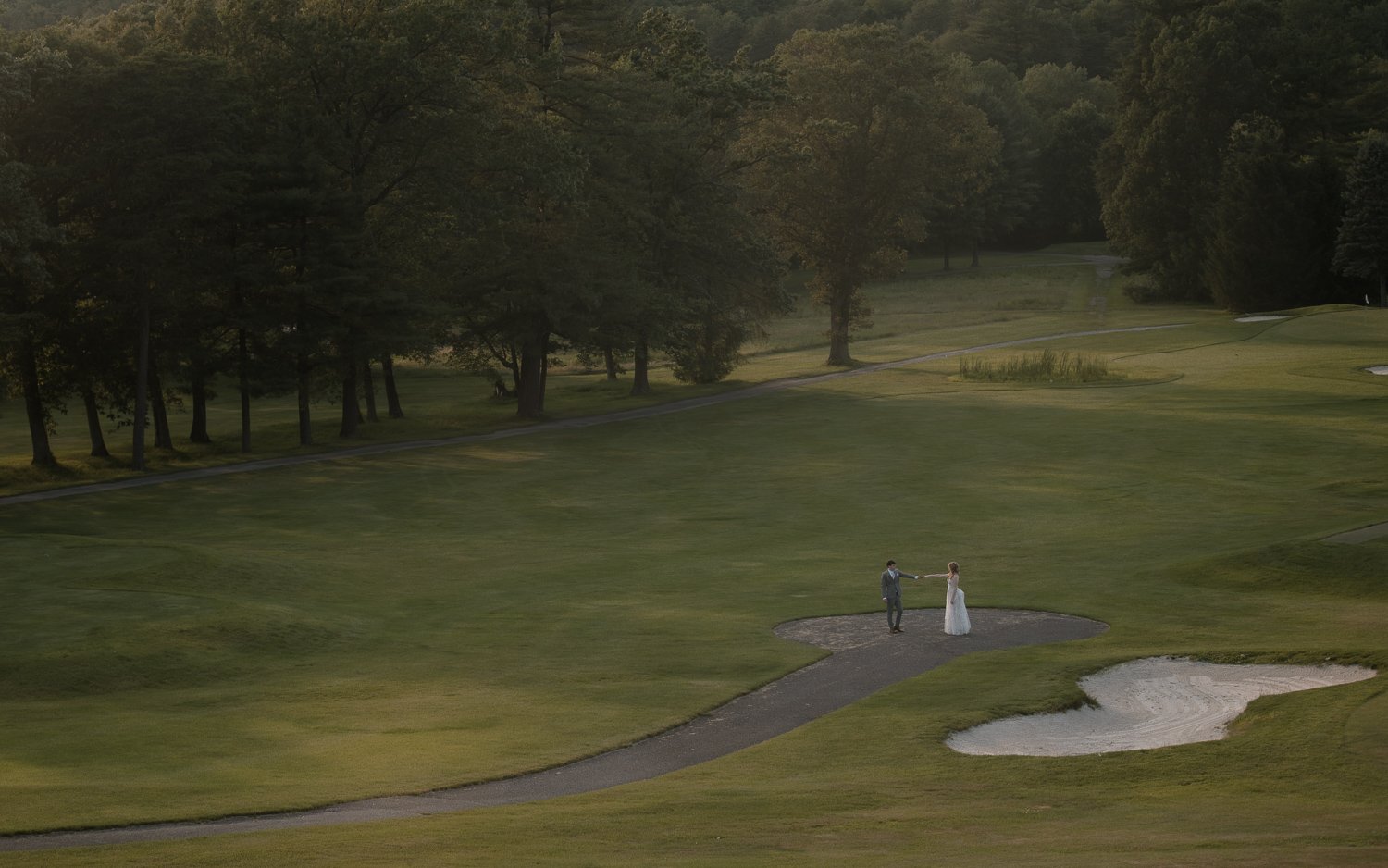 A bride and groom stand on a small dirt patch near a golf course, holding hands and facing each other in a scenic outdoor setting with trees, grass, and a sand trap.