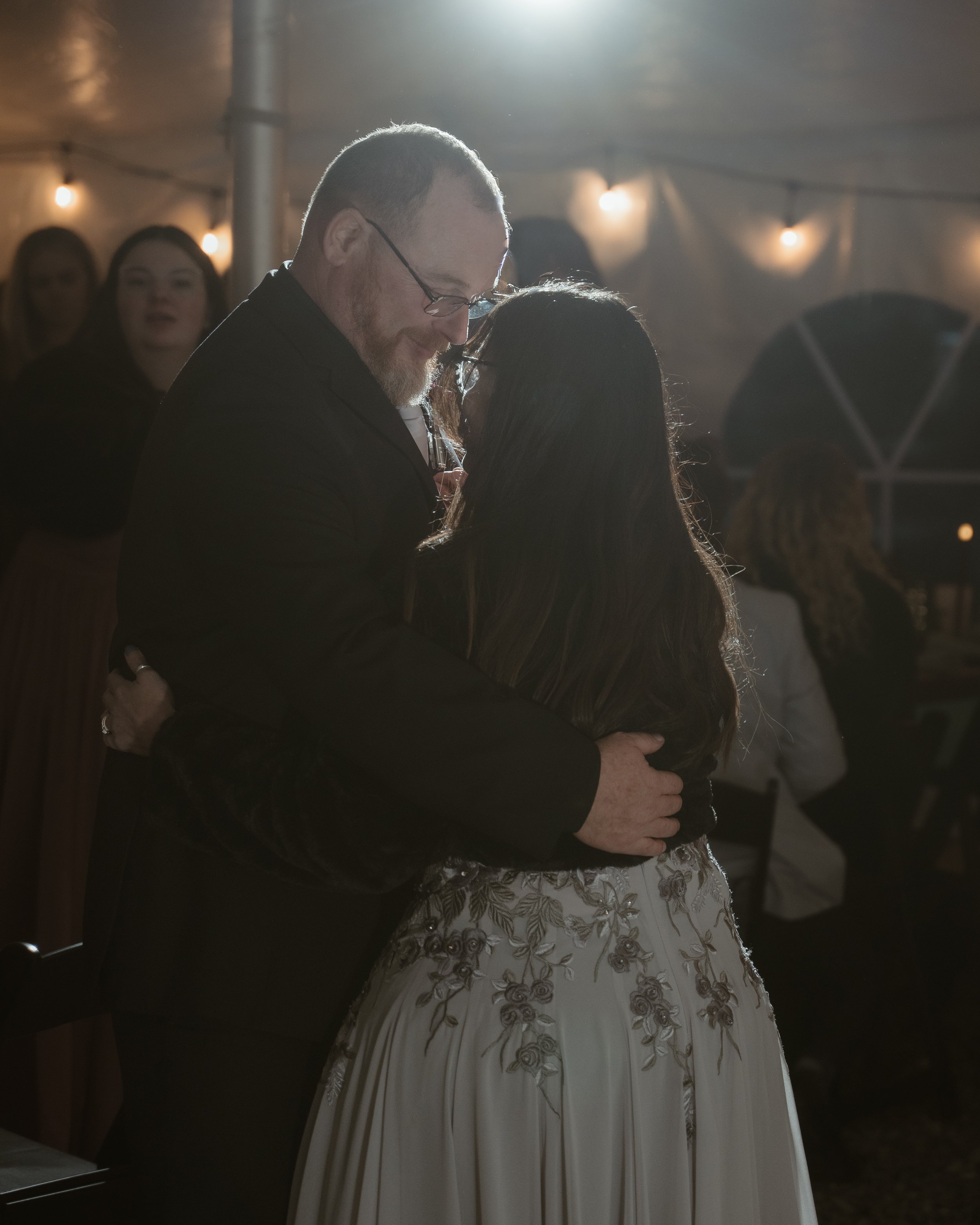 A man and woman sharing an intimate dance at a celebration or wedding reception, with warm lighting and guests in the background.