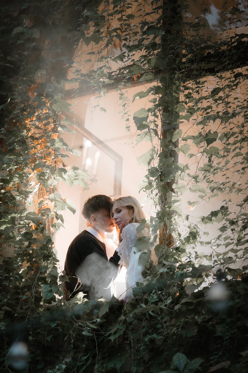 A couple stands close together amid lush green foliage, with sunlight streaming in from behind, creating a dreamy and intimate atmosphere.