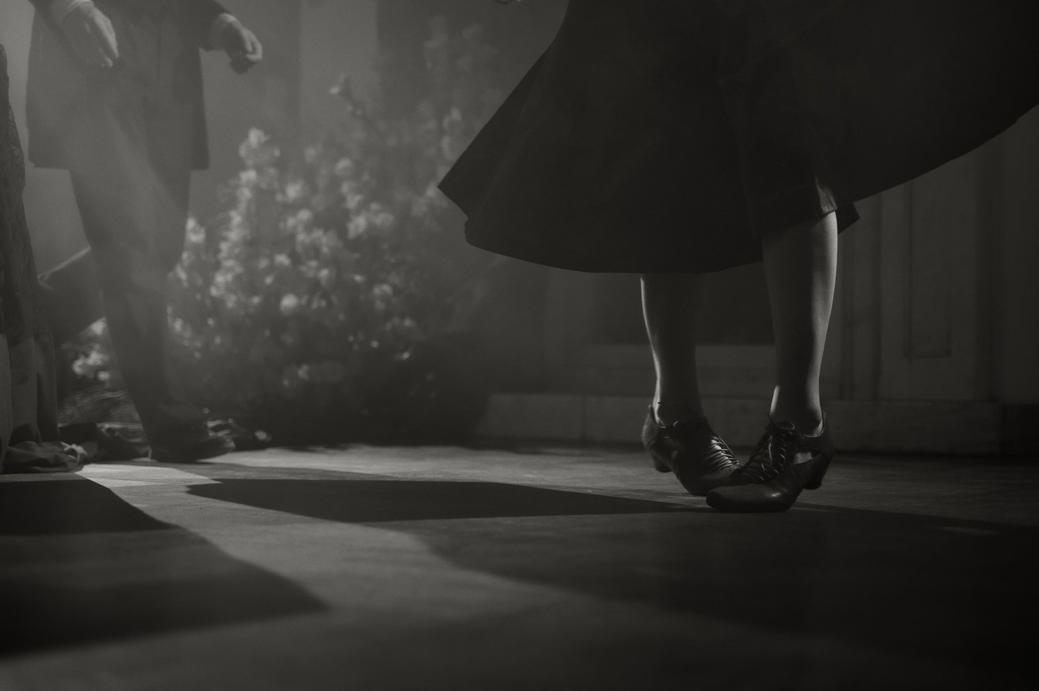 A person in a floral skirt and tights is dancing on a wooden floor, with their feet in a dance position. The photo is in black and white and shows a blurred figure in the background, possibly a man, with what appears to be a garden scene outside the window.