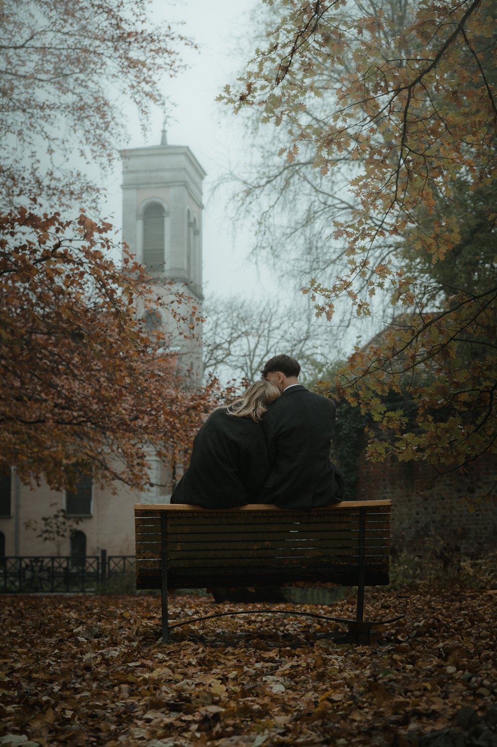 A couple sitting close together on a park bench in autumn, surrounded by falling leaves and orange trees, with a church tower in the background.