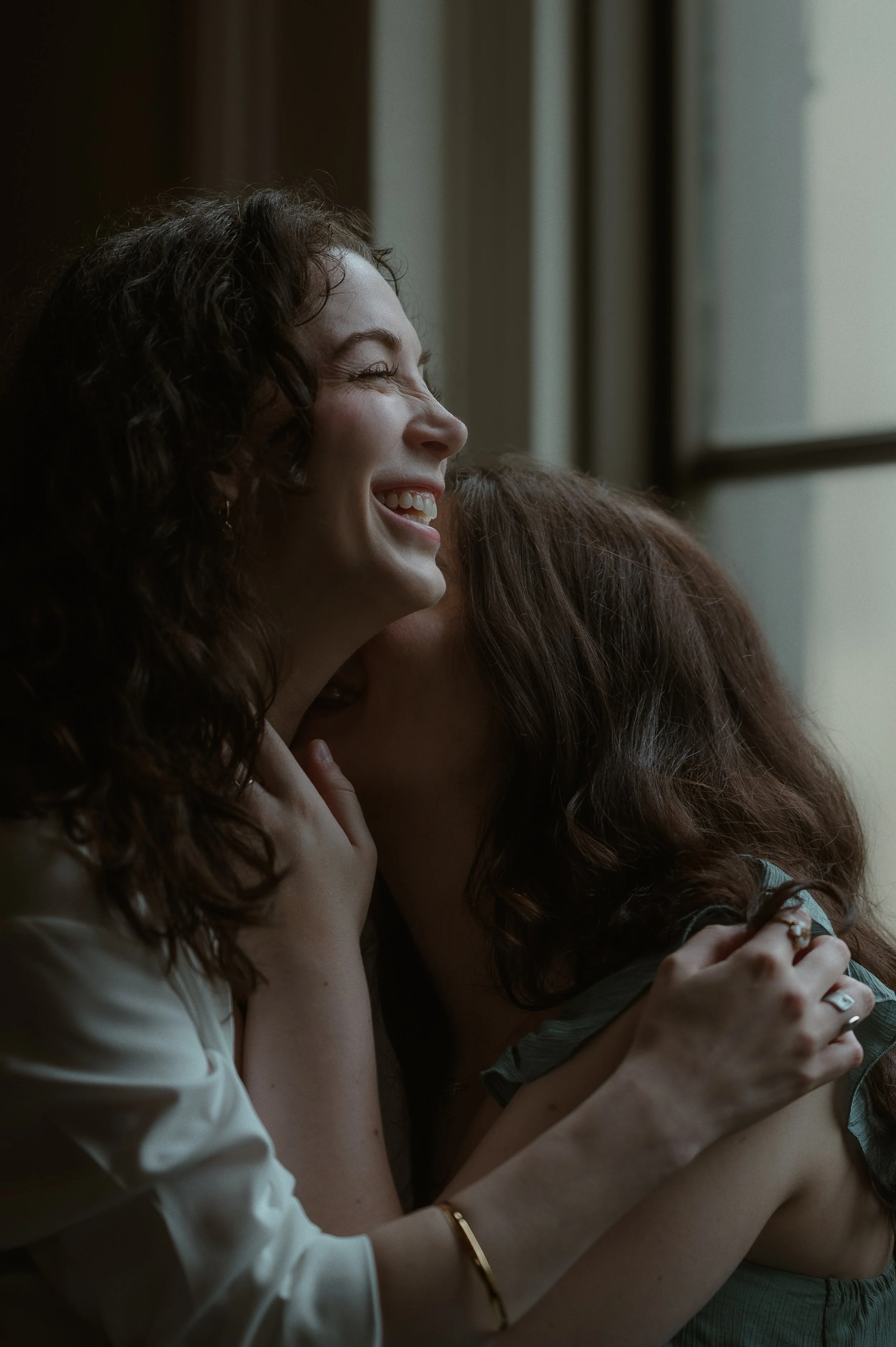 Two women sharing a joyful, intimate moment, hugging and smiling near a window.