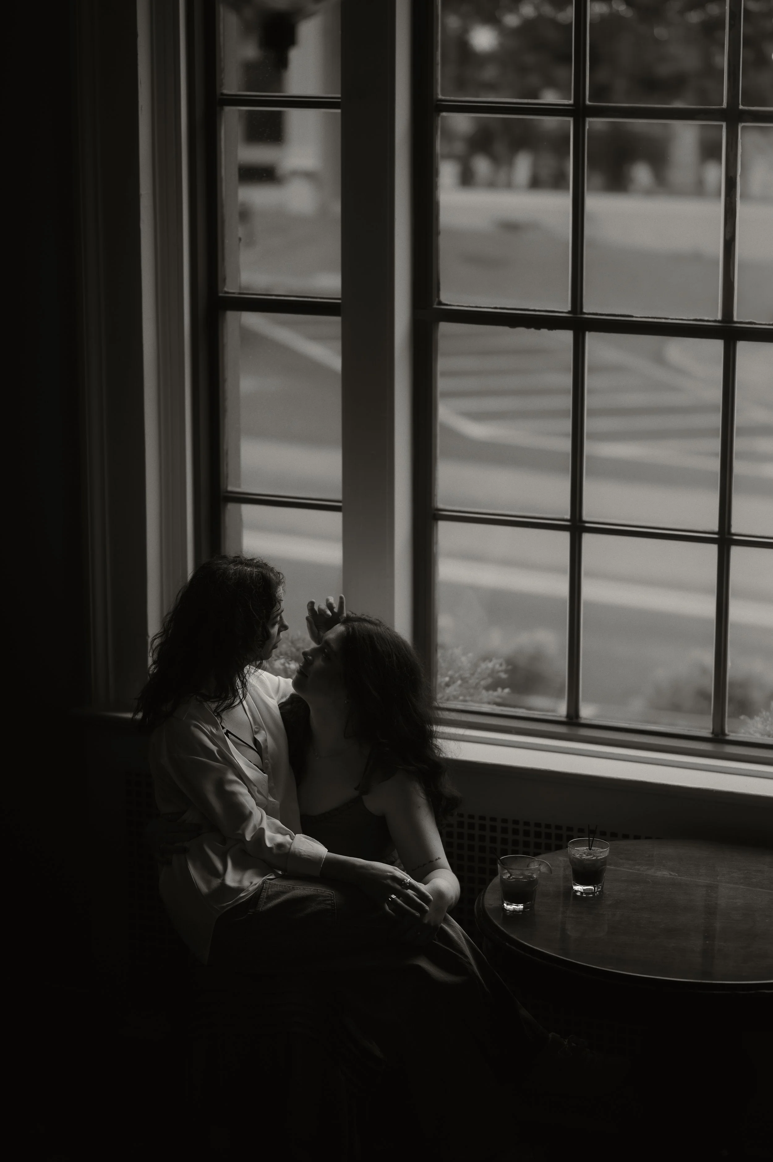 Two women sitting by a large window, sharing an intimate moment, with drinks on a small round table nearby, in a dimly lit setting.