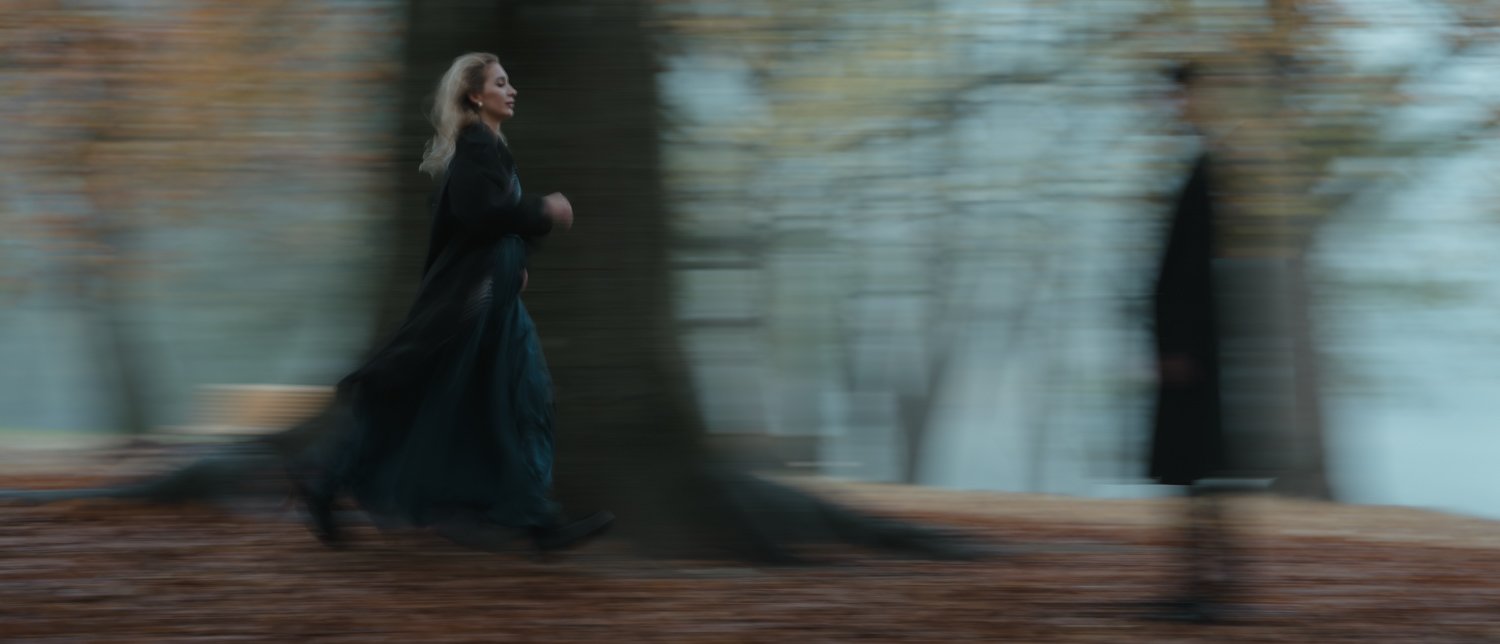 A woman with blonde hair running through a forest with a blurred man standing near a tree in the background.