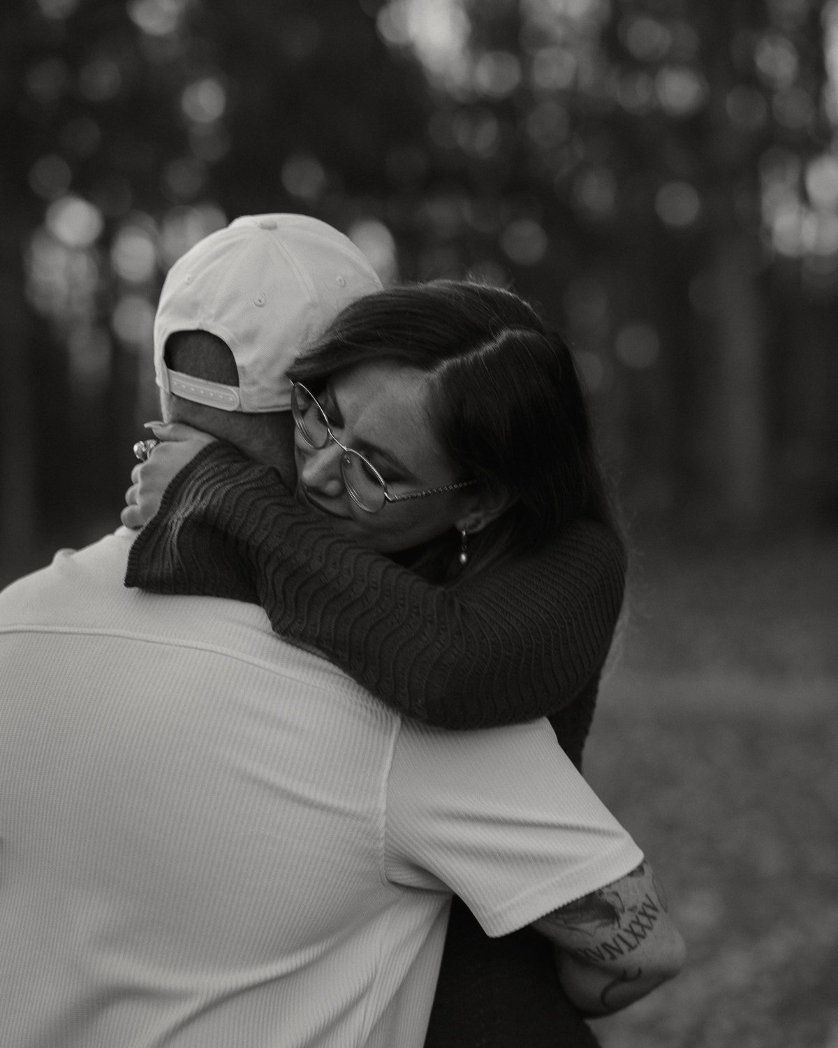 A woman embraces a man who is wearing a baseball cap, with her eyes closed, in an outdoor setting with blurred trees in the background.