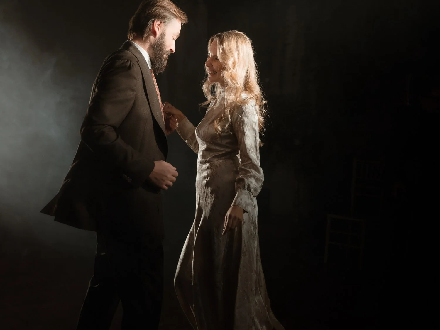 A man and woman standing close, smiling and looking at each other in a dimly lit room.