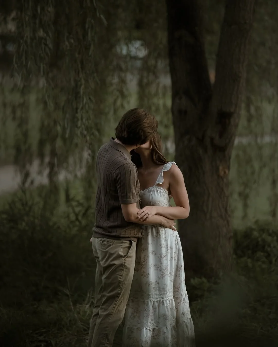 A couple stands close together outdoors, embracing and kissing near a large tree surrounded by greenery.