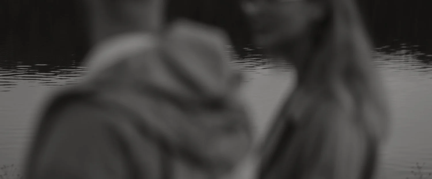 Two people are seen at the edge of a body of water, with their silhouettes blurred, possibly taking a selfie or sharing a moment.