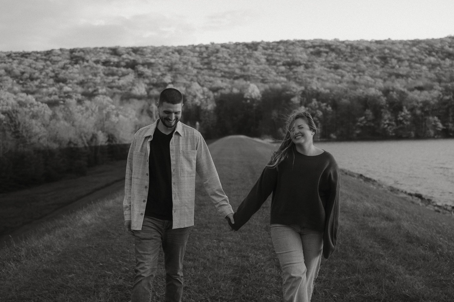 A black and white photo of a smiling couple walking hand in hand on a grassy path near a body of water with trees and hills in the background.