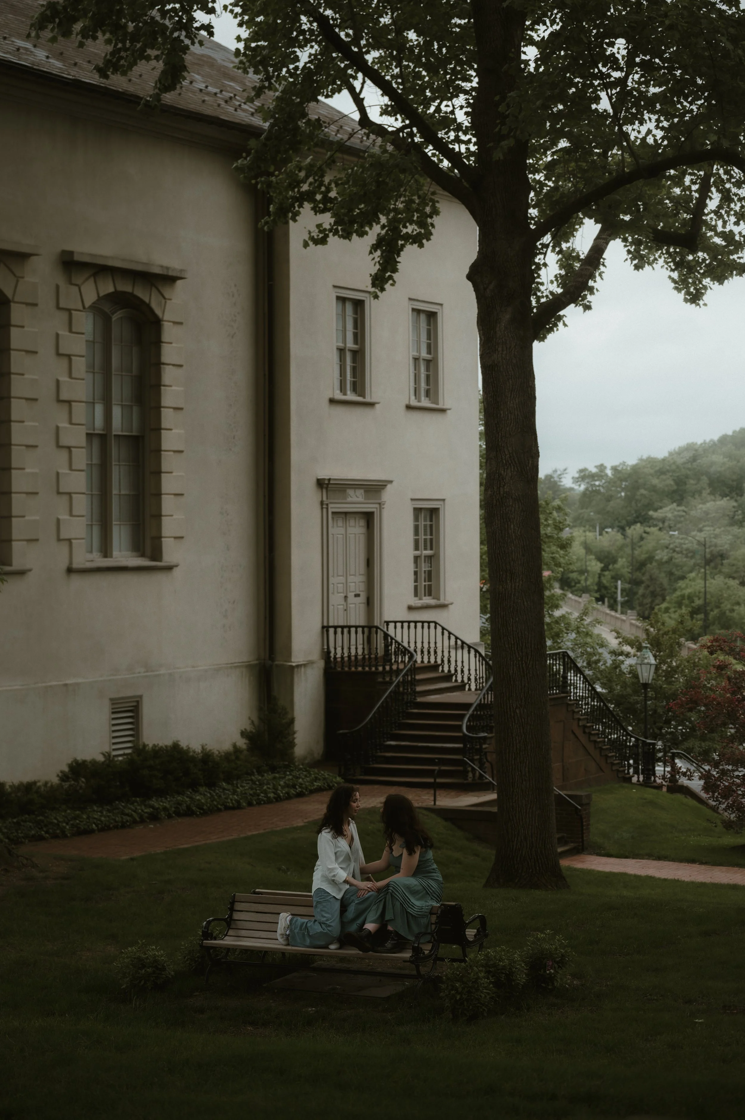 Two women sit closely on a park bench underneath a large tree, near a historic residential building with a staircase and multiple windows.