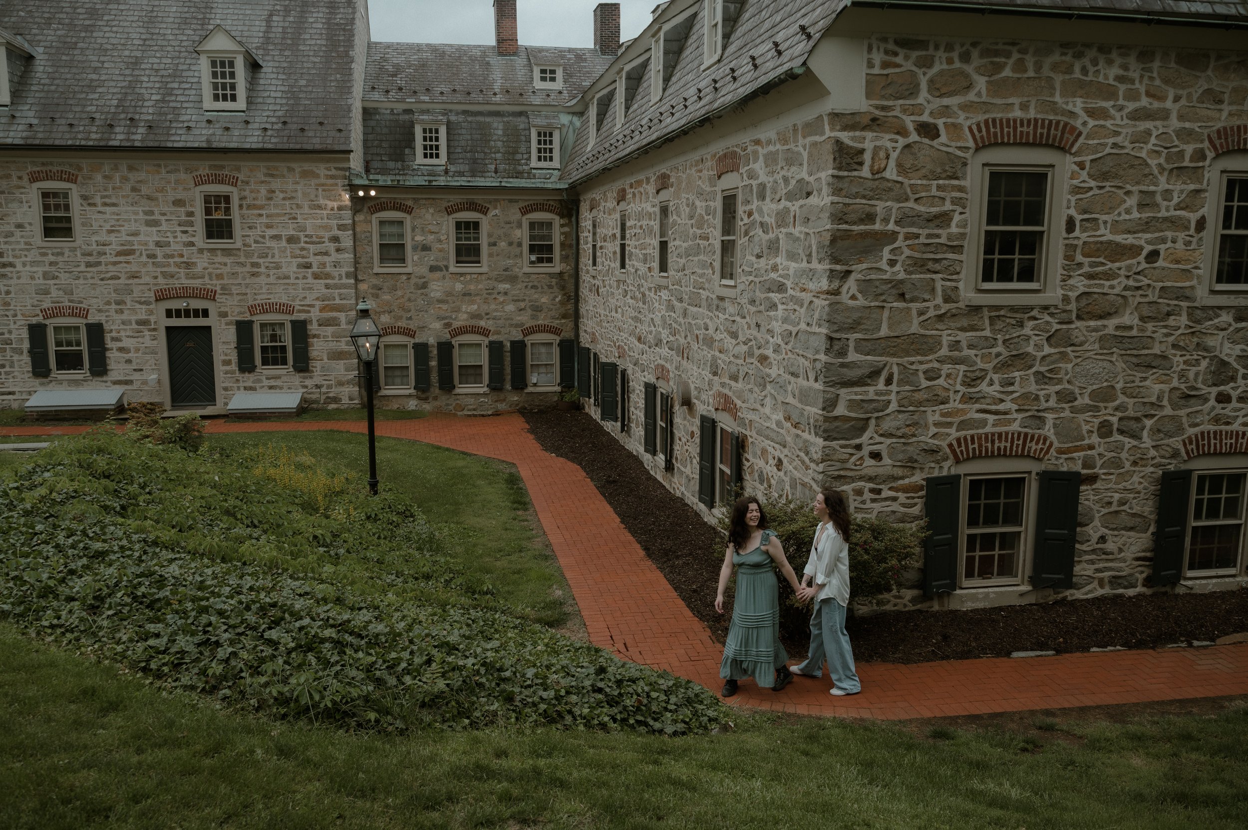 Two women holding hands and smiling on a brick pathway in front of a stone building with multiple windows and black shutters, during daytime.