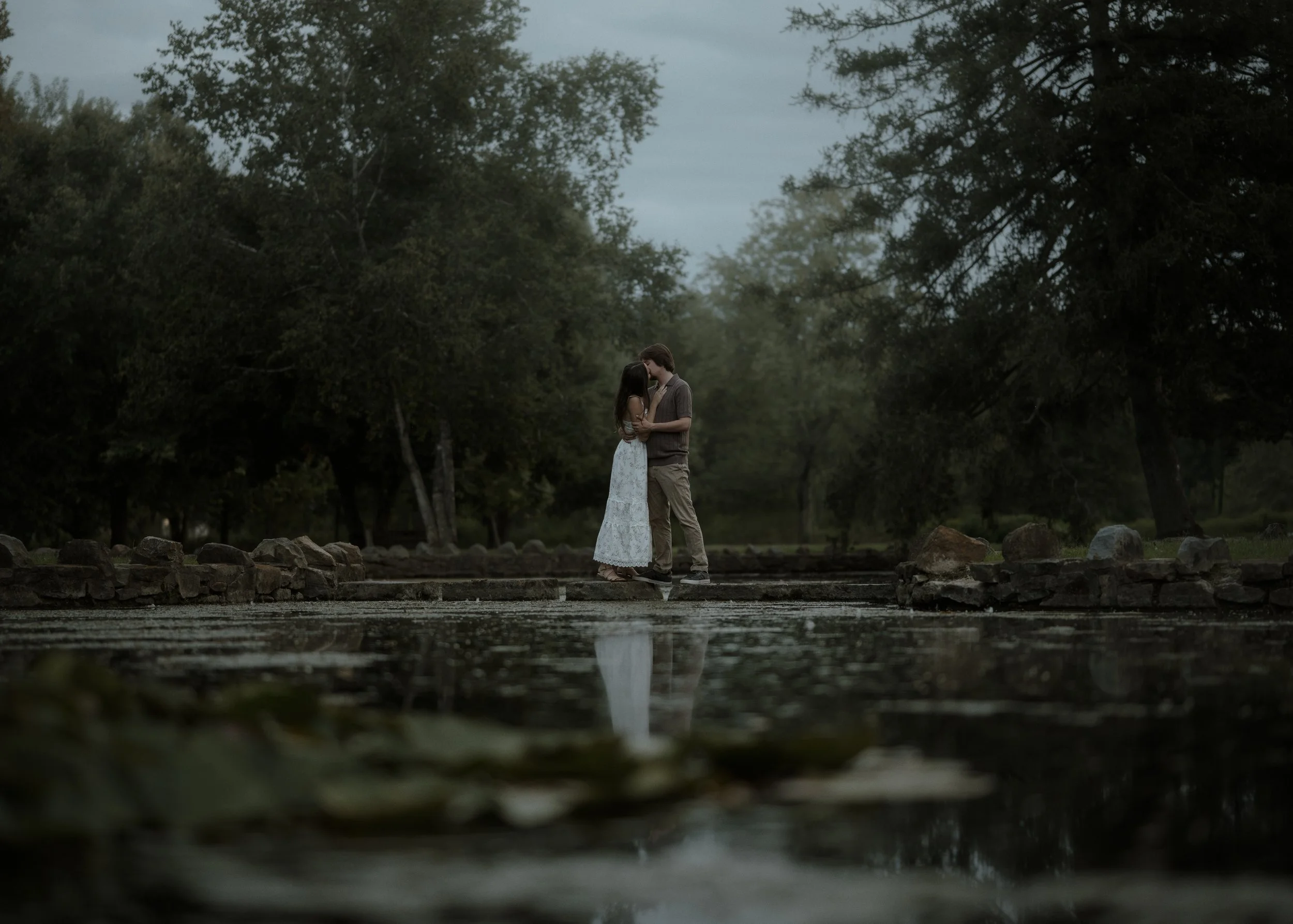 A young couple standing on a rocky path by a pond, embracing and about to kiss, surrounded by trees, during twilight or early evening.