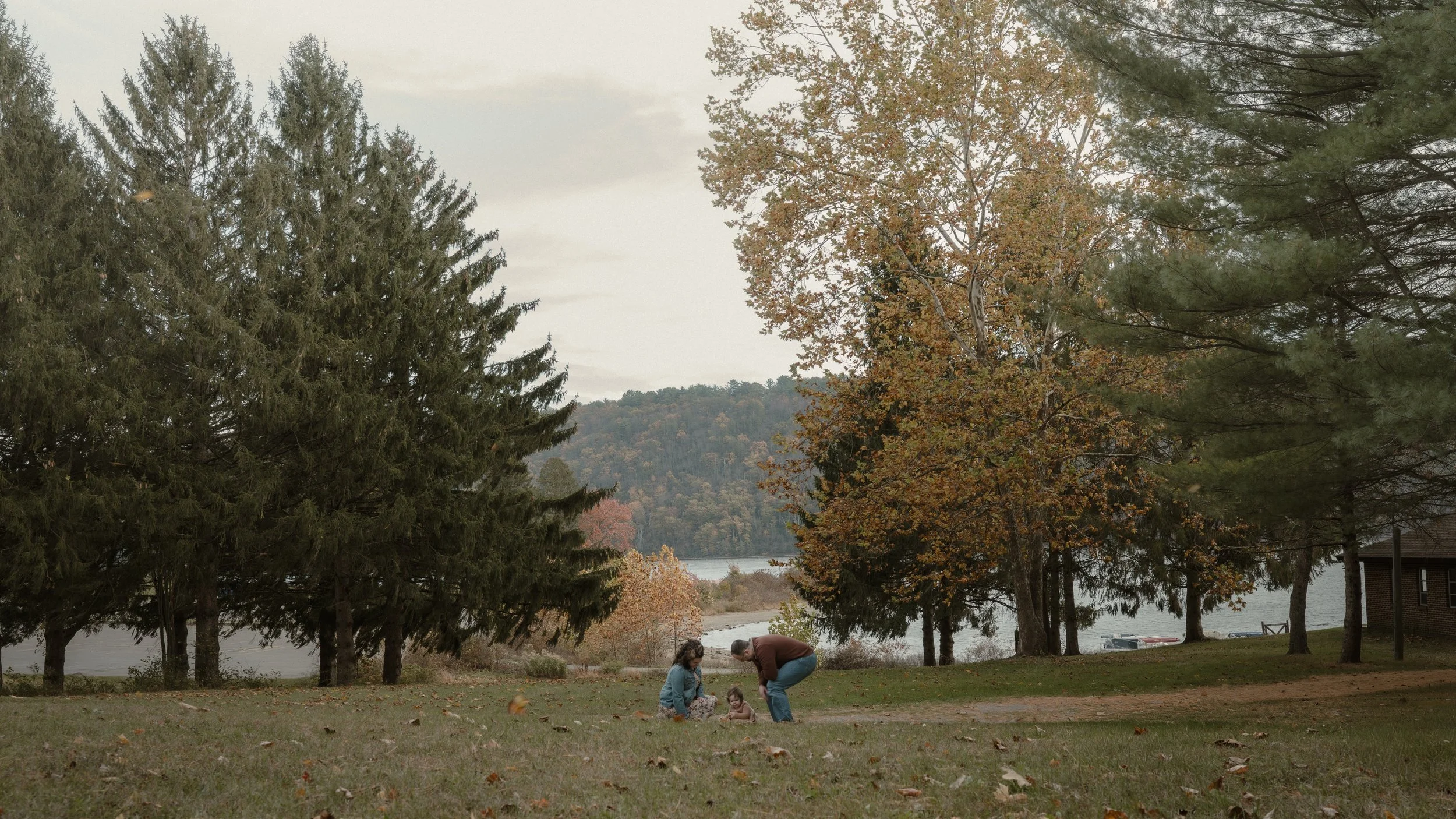 Family with two children playing on a grassy lawn near a lake, surrounded by trees with fall foliage.