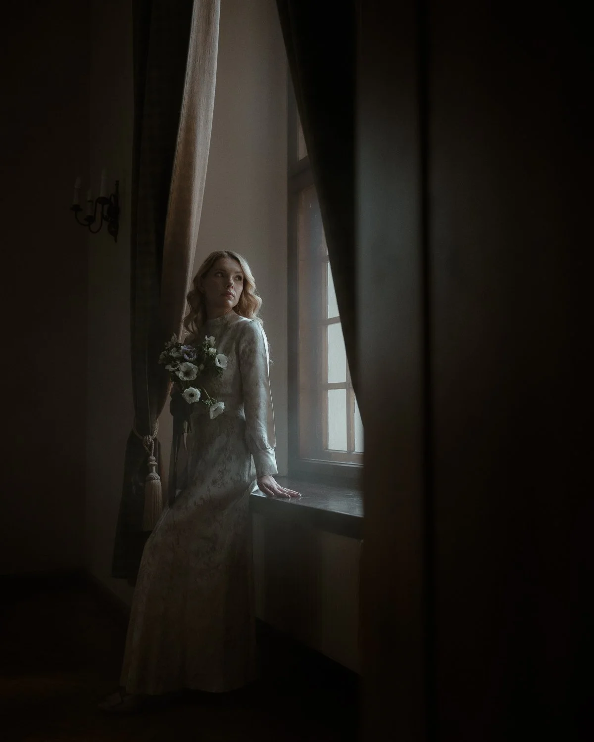A woman in a vintage-style dress holding a bouquet, leaning on a windowsill and looking outside through a window, partially obscured by curtains.