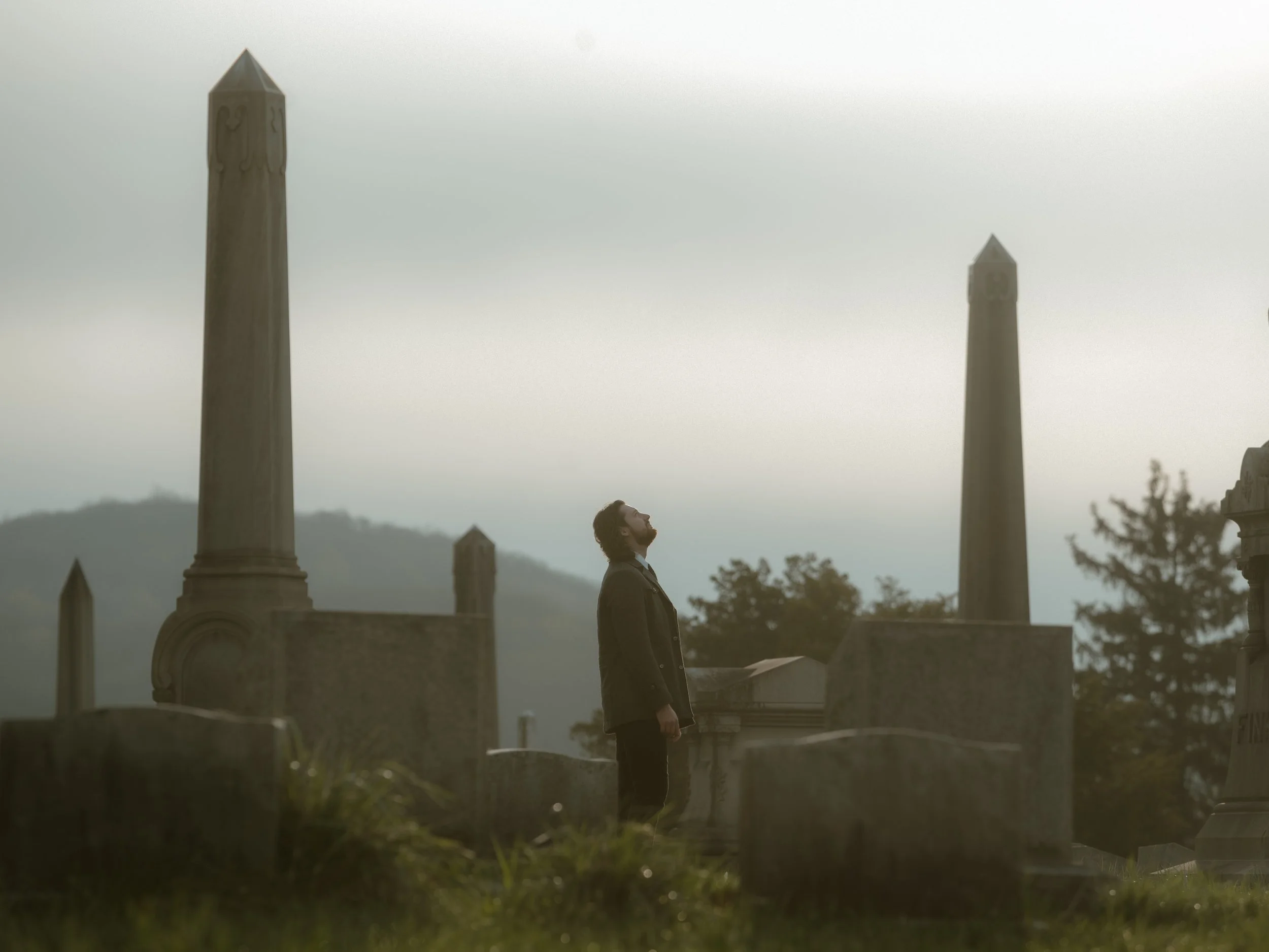 A man in a coat standing in a cemetery with tombstones and tall obelisks, looking upward, with hills and cloudy sky in the background.