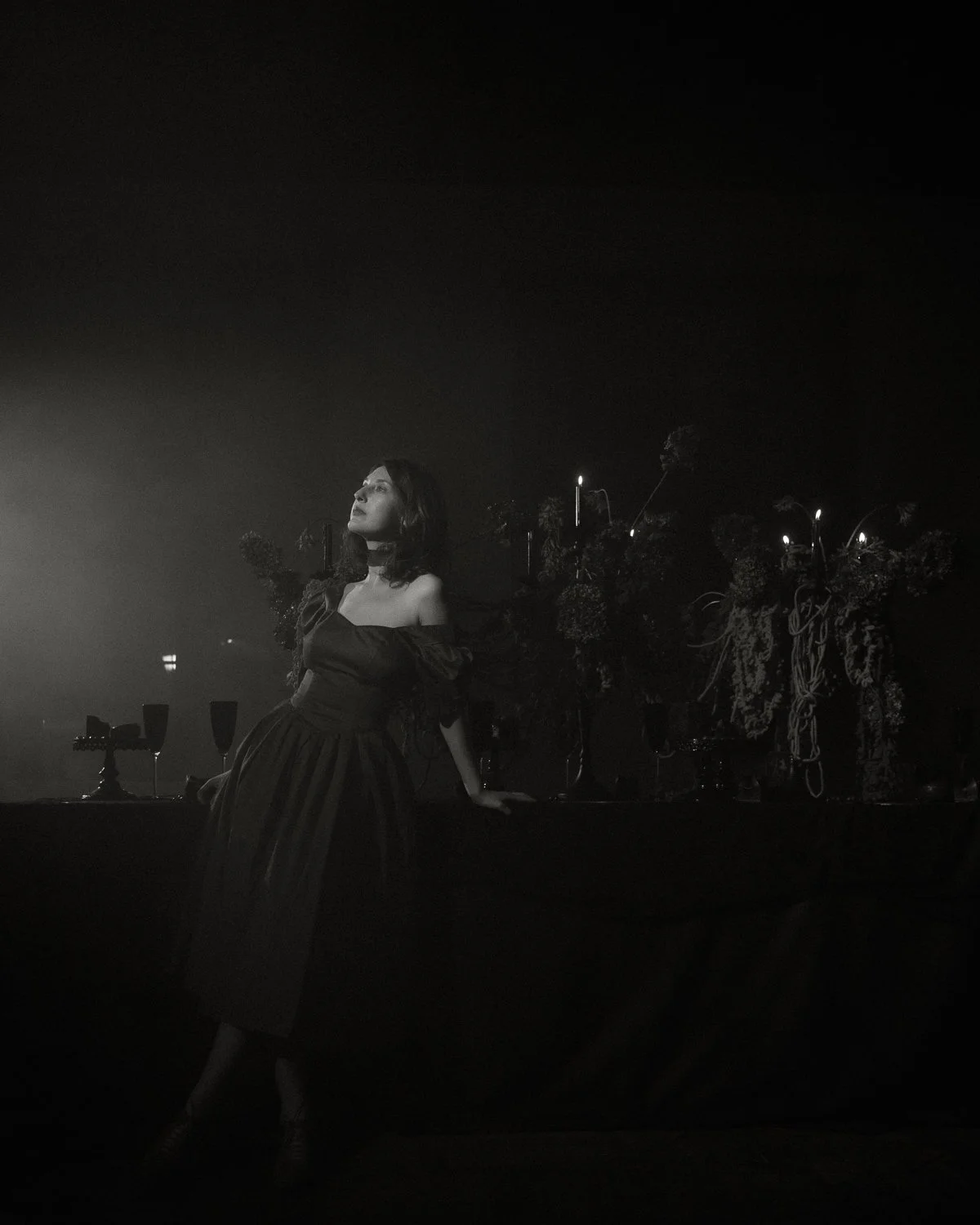 A woman in a dark dress with off-the-shoulder sleeves is standing against a dark background, with some decorative elements and candles behind her, illuminated by a soft light.