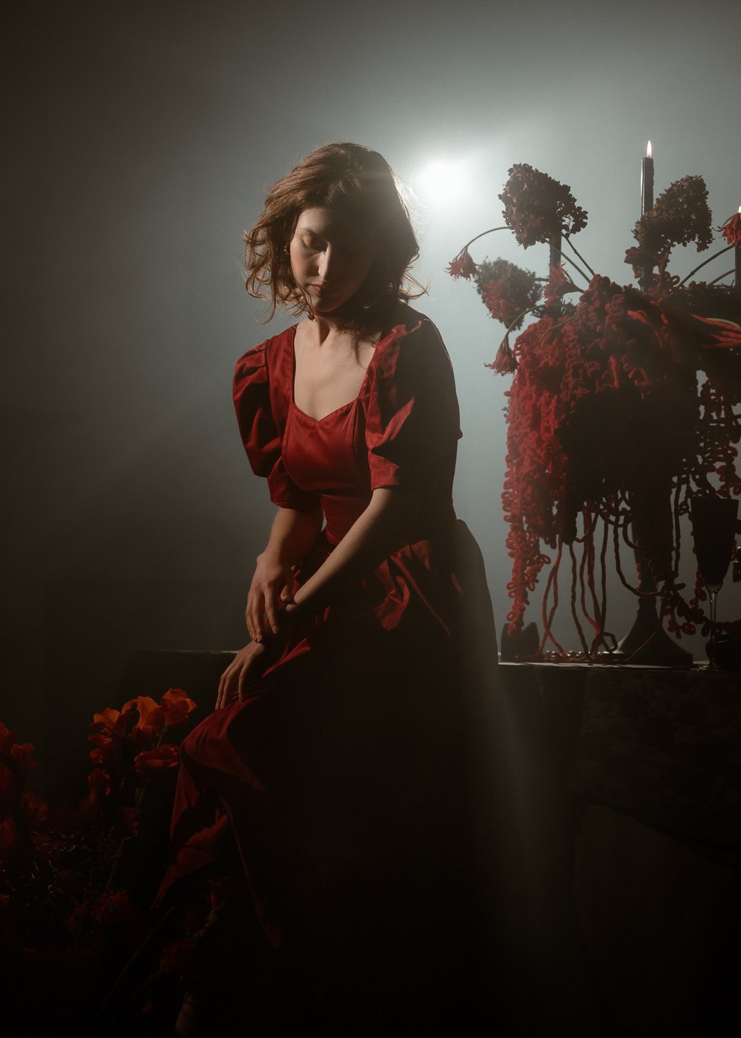 A woman in a red dress with puffy sleeves sitting beside a table with a large floral arrangement and a lit candle, dimly lit with a haze and backlighting