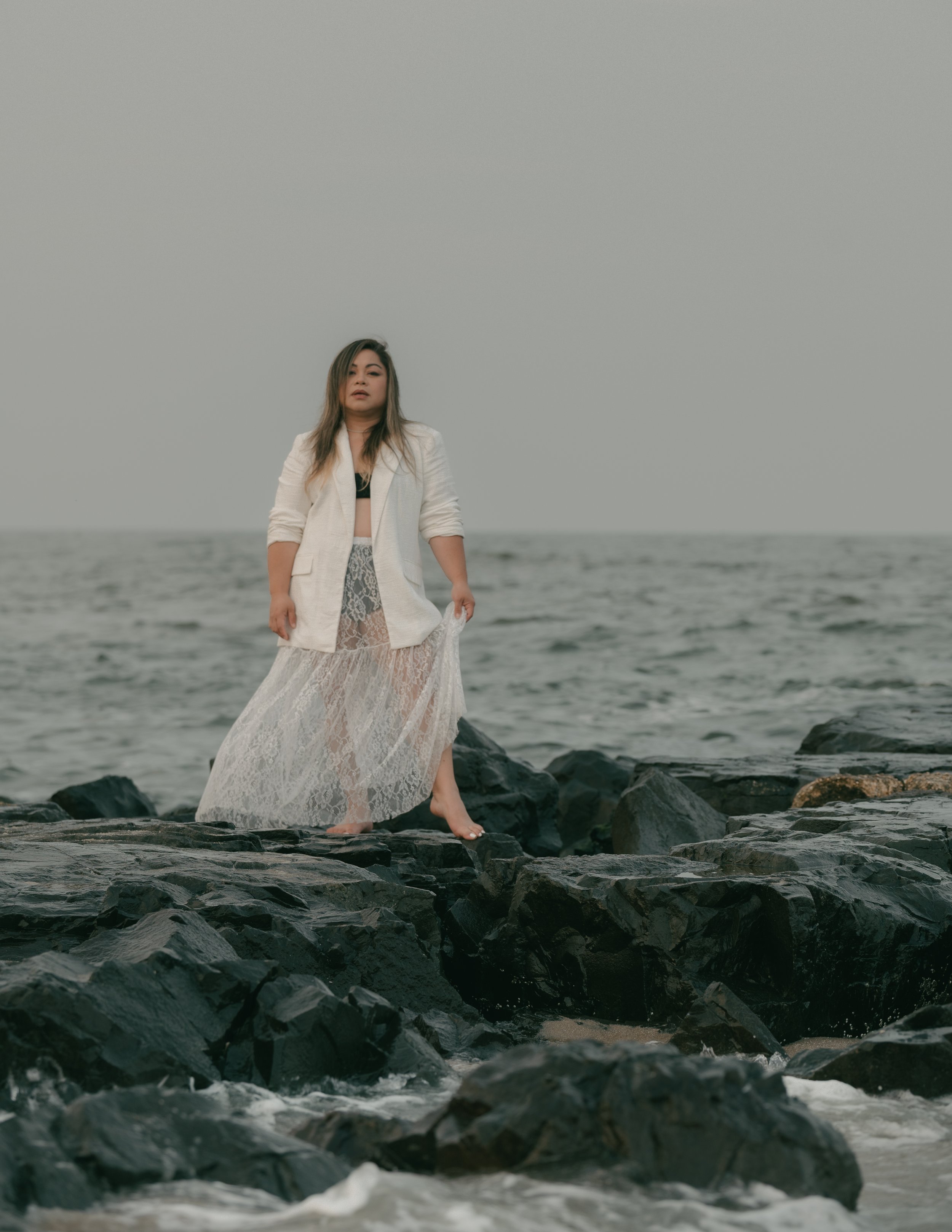 Woman standing barefoot on rocks at the beach, wearing a white blazer and a long lace skirt, with the ocean in the background.