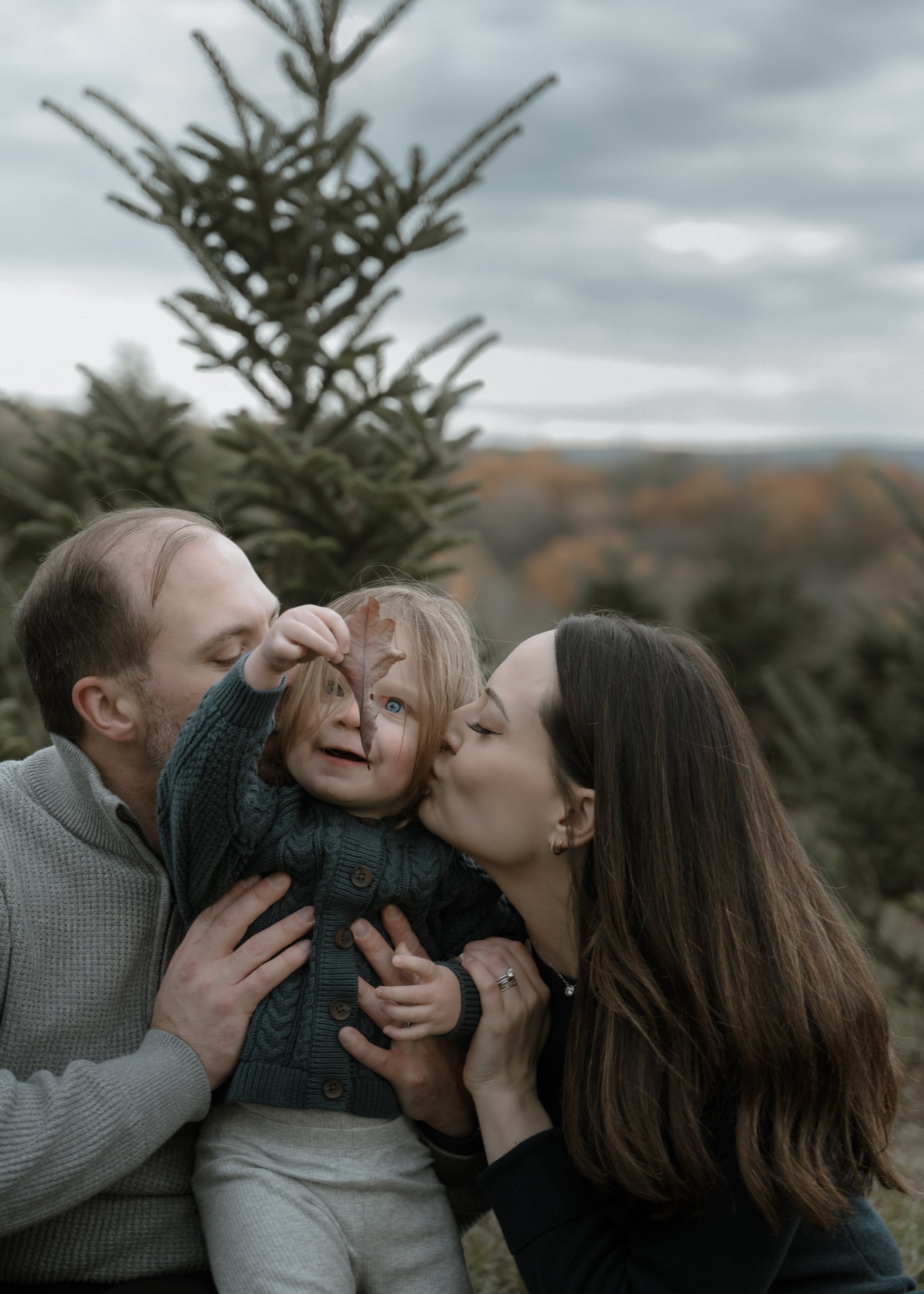 Family of three outdoors: father, mother, and young daughter enjoys moment with daughter holding a leaf, mother kisses daughter, father holds daughter against backdrop of trees and cloudy sky.