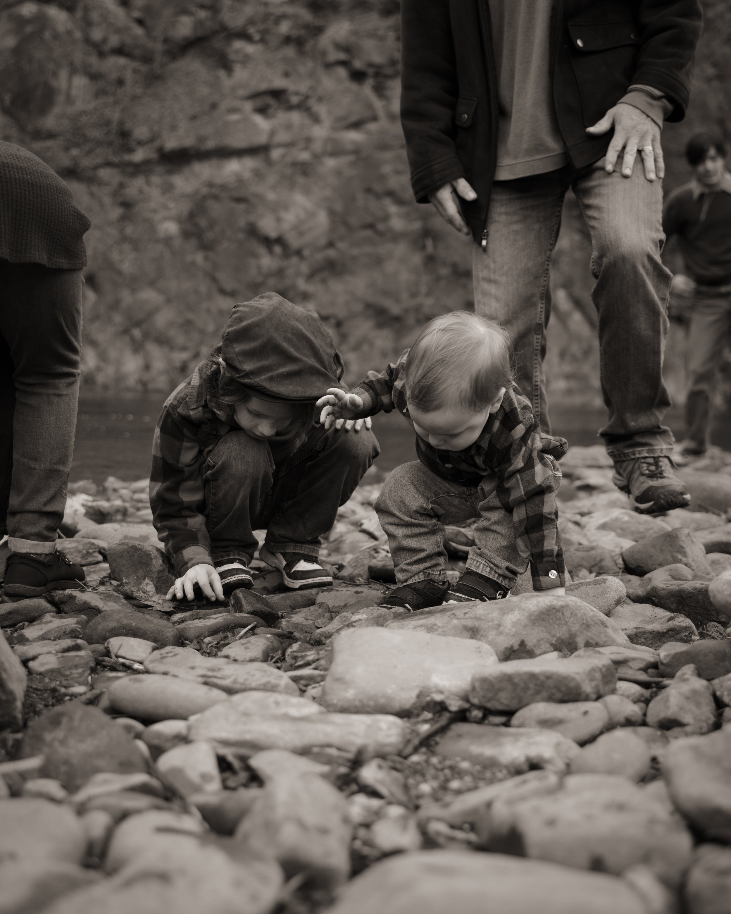 Two young children crouching on a rocky riverbank, exploring the rocks, with an adult standing nearby and other children in the background.