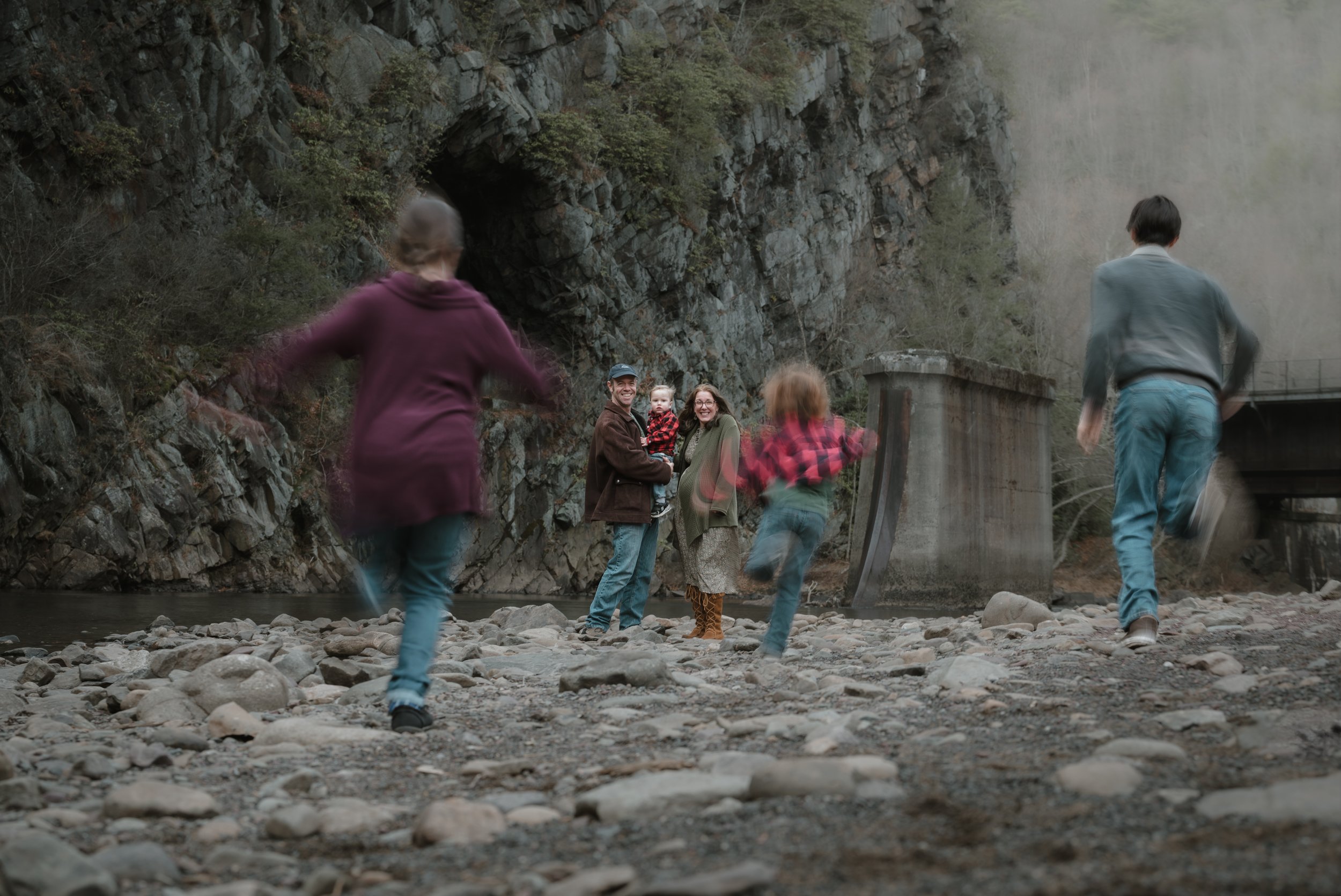 Family enjoying outdoors near a rocky lakeshore under a bridge, with children running and a couple standing with a toddler.