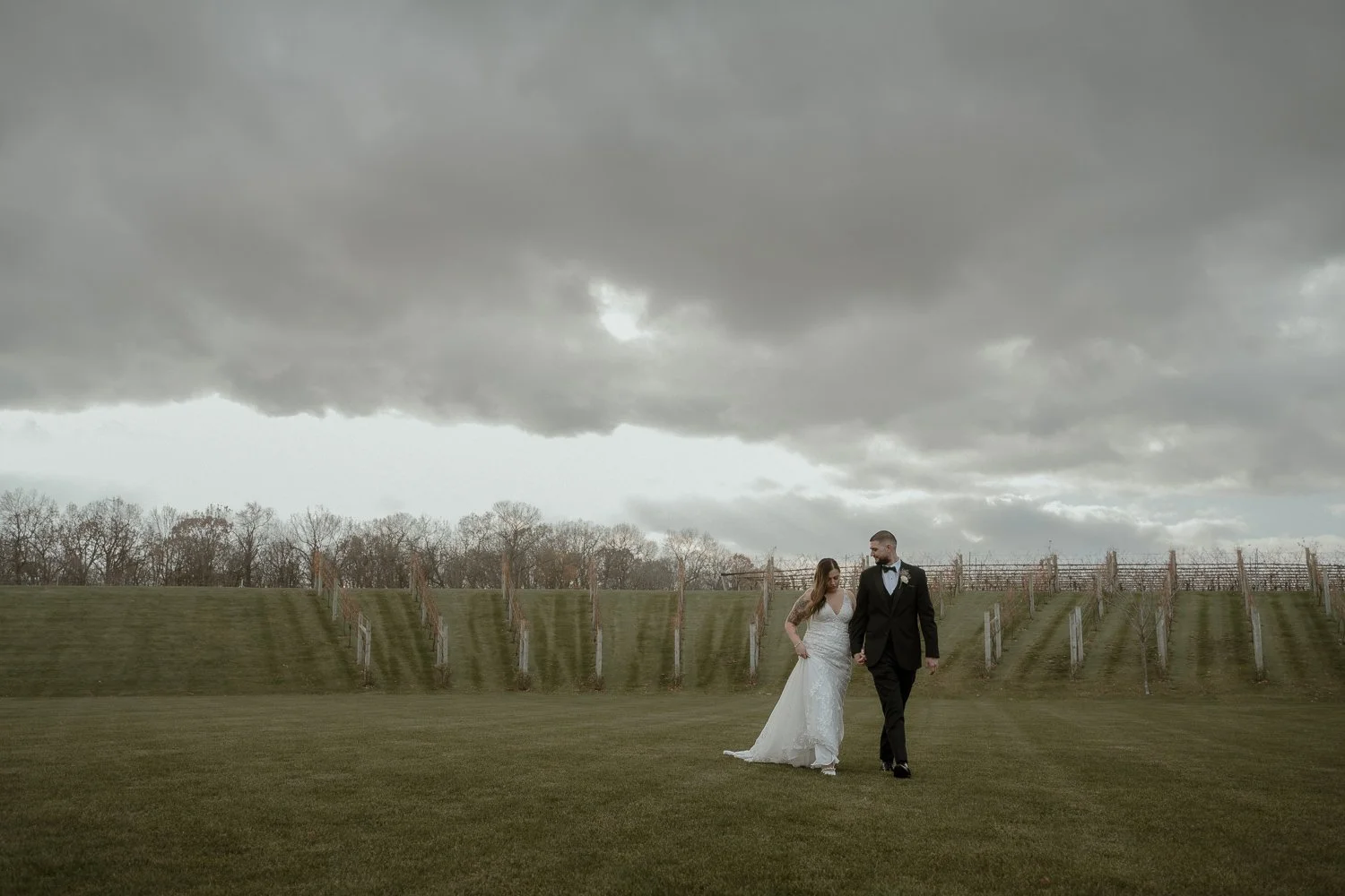 A bride in a white wedding dress and a groom in a black tuxedo walking hand in hand on a grassy field with a cloudy sky overhead.
