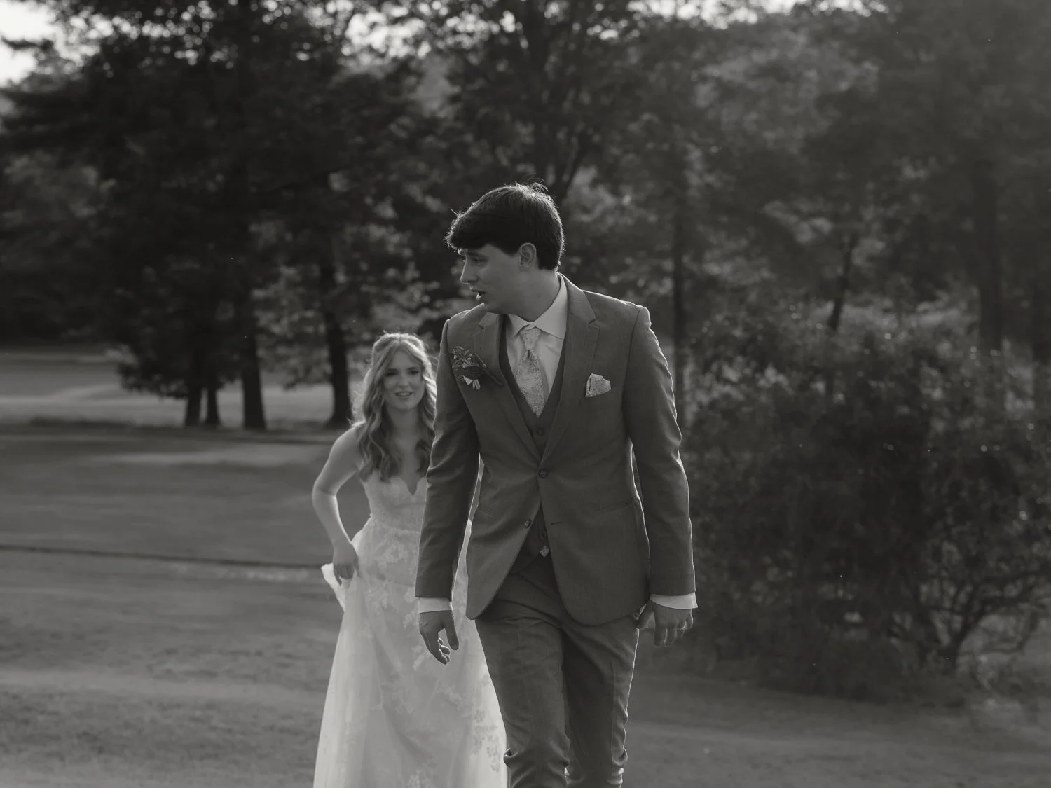 A black and white photo of a bride and groom walking outdoors, with the bride smiling and the groom looking off to the side, surrounded by trees and grass in the background.