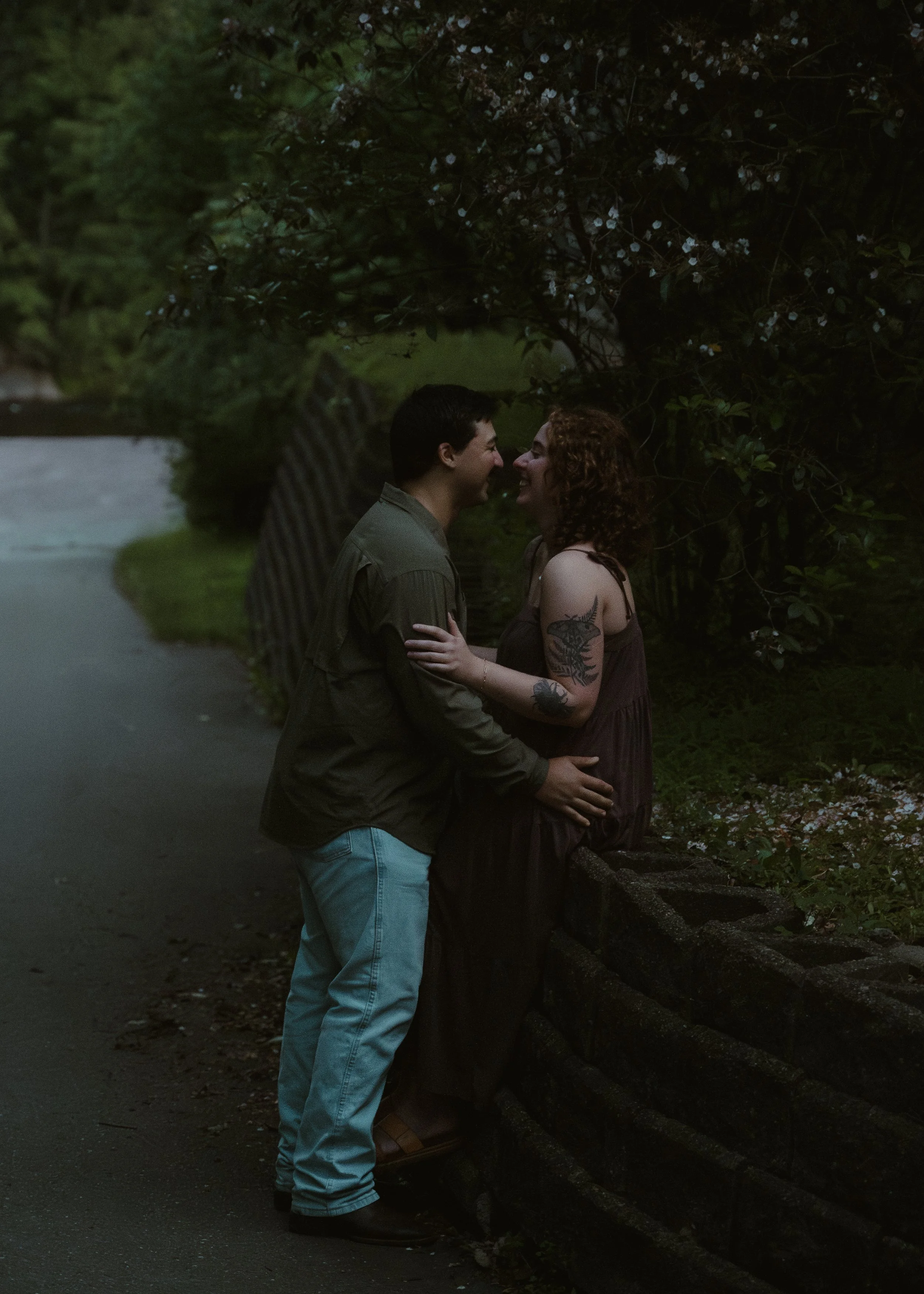 A couple standing close together by a stone wall, smiling and touching foreheads in an outdoor setting with trees and flowers.