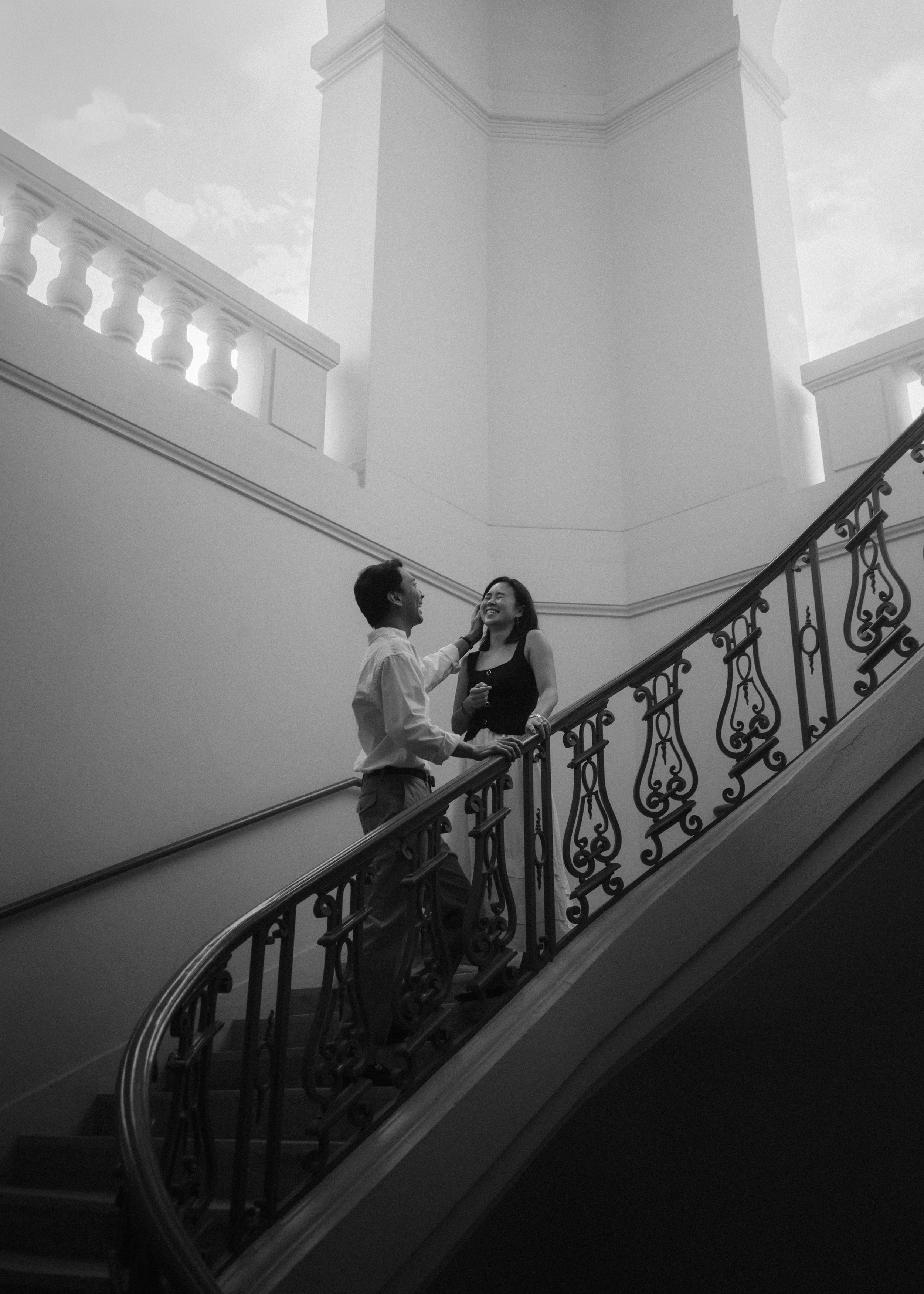 A man and woman standing on a staircase, smiling and engaging in conversation inside a building with classical architectural details.
