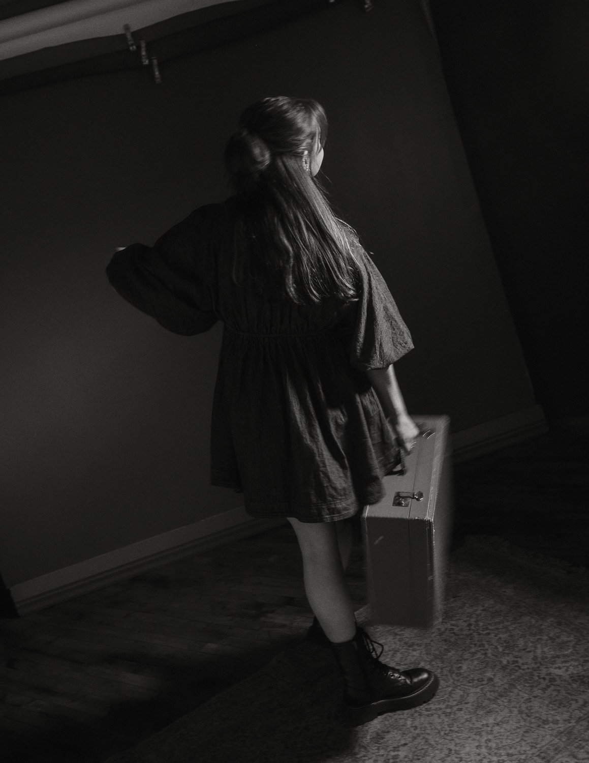 A girl with long hair, dressed in a black dress and combat boots, standing indoors on a hardwood floor with a suitcase.