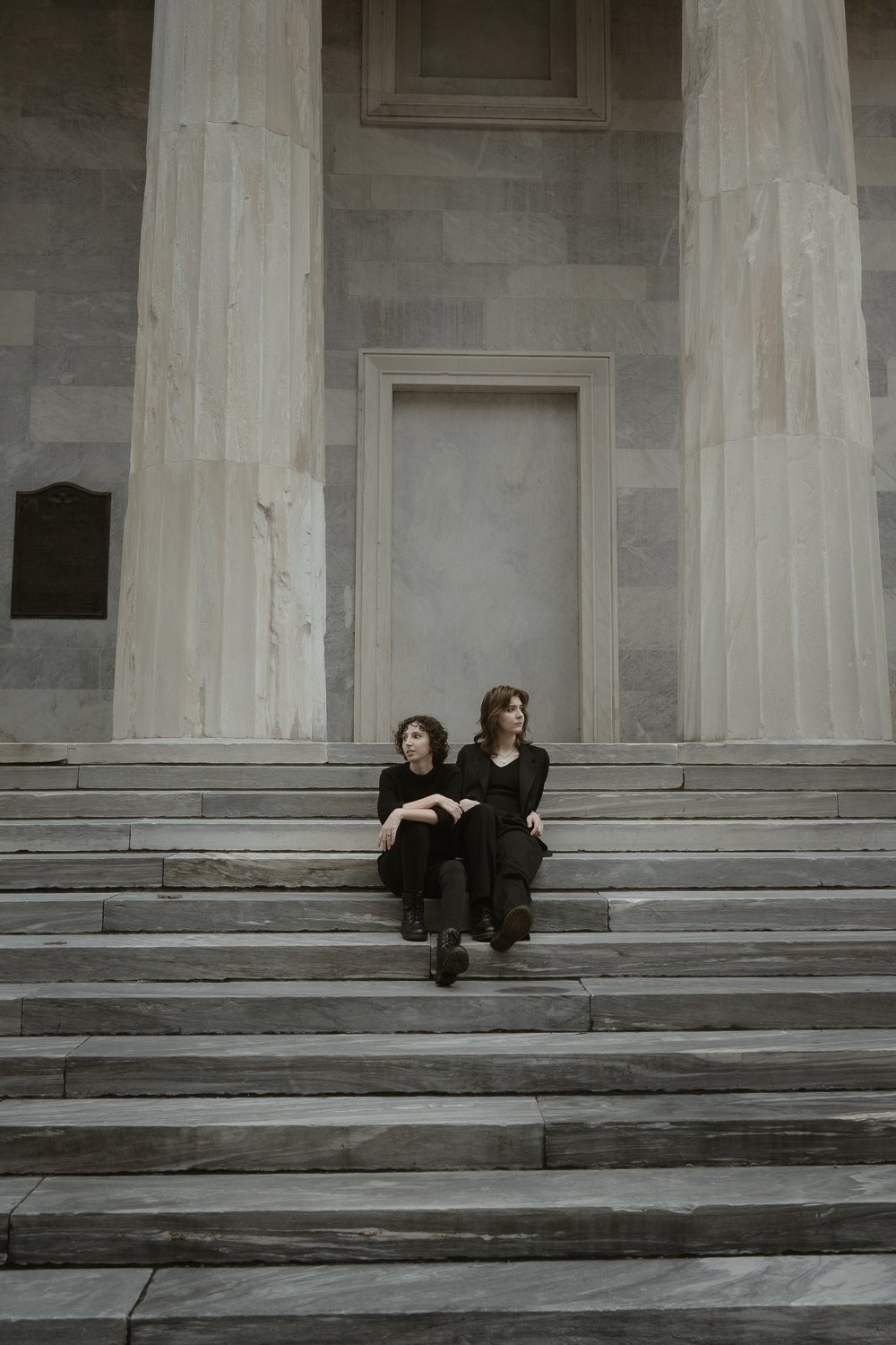 Two women sitting on the steps of a large, classical stone building with tall columns and a door behind them, dressed in black clothing.