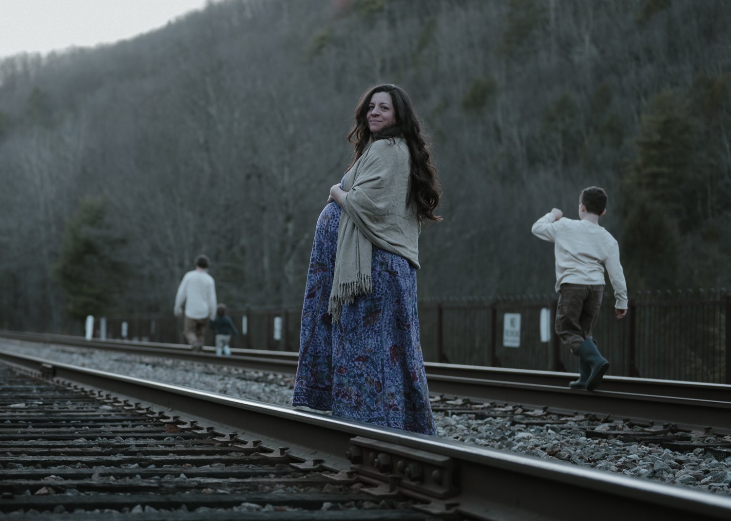 A woman in a long blue patterned skirt and beige shawl standing on train tracks. In the background, a boy in a white shirt and boots is walking along the tracks, and another person is farther ahead, walking away.