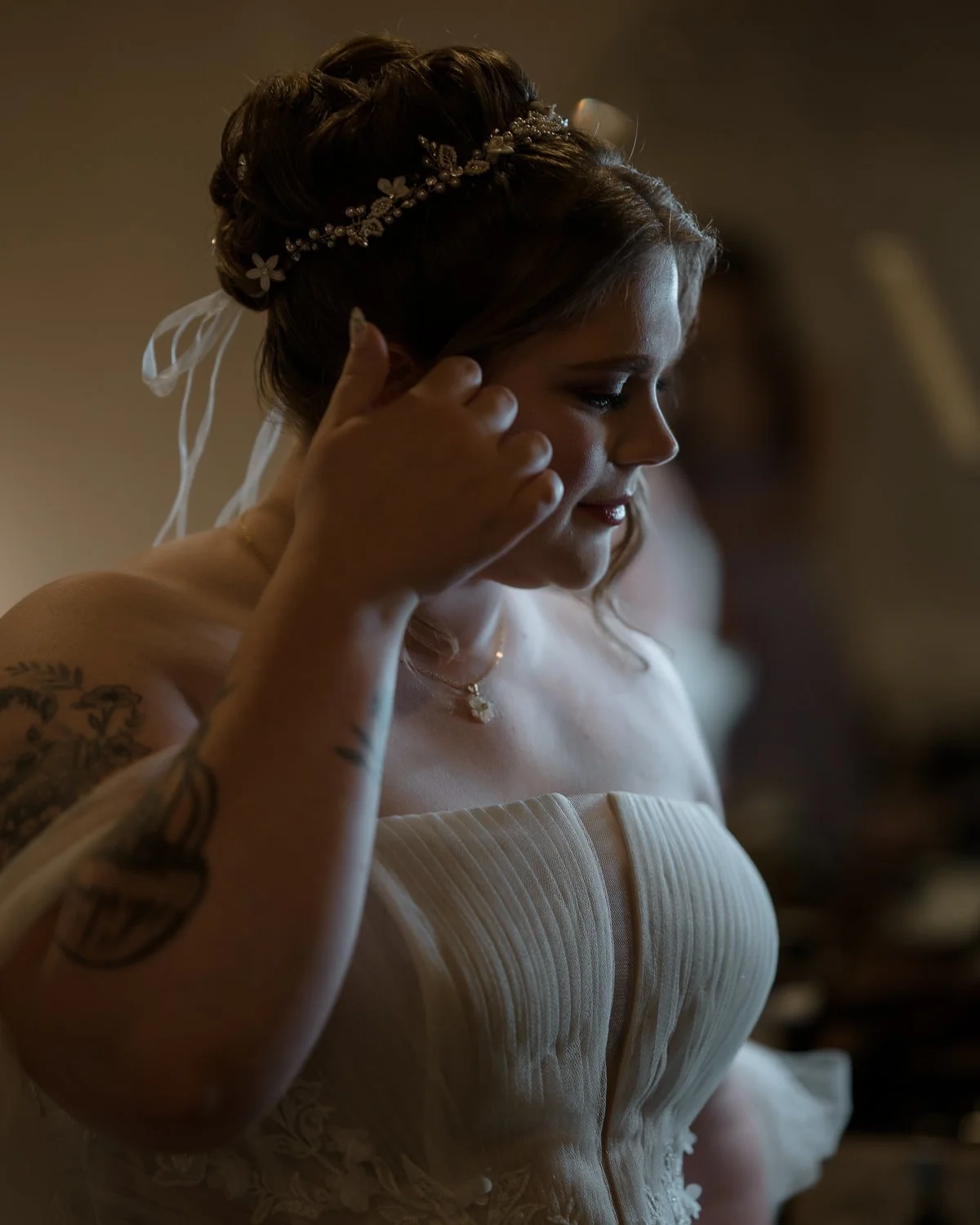 A bride in a strapless wedding dress with styled hair wearing a decorative headband, being helped with her earring by a person with tattoos on their arm.