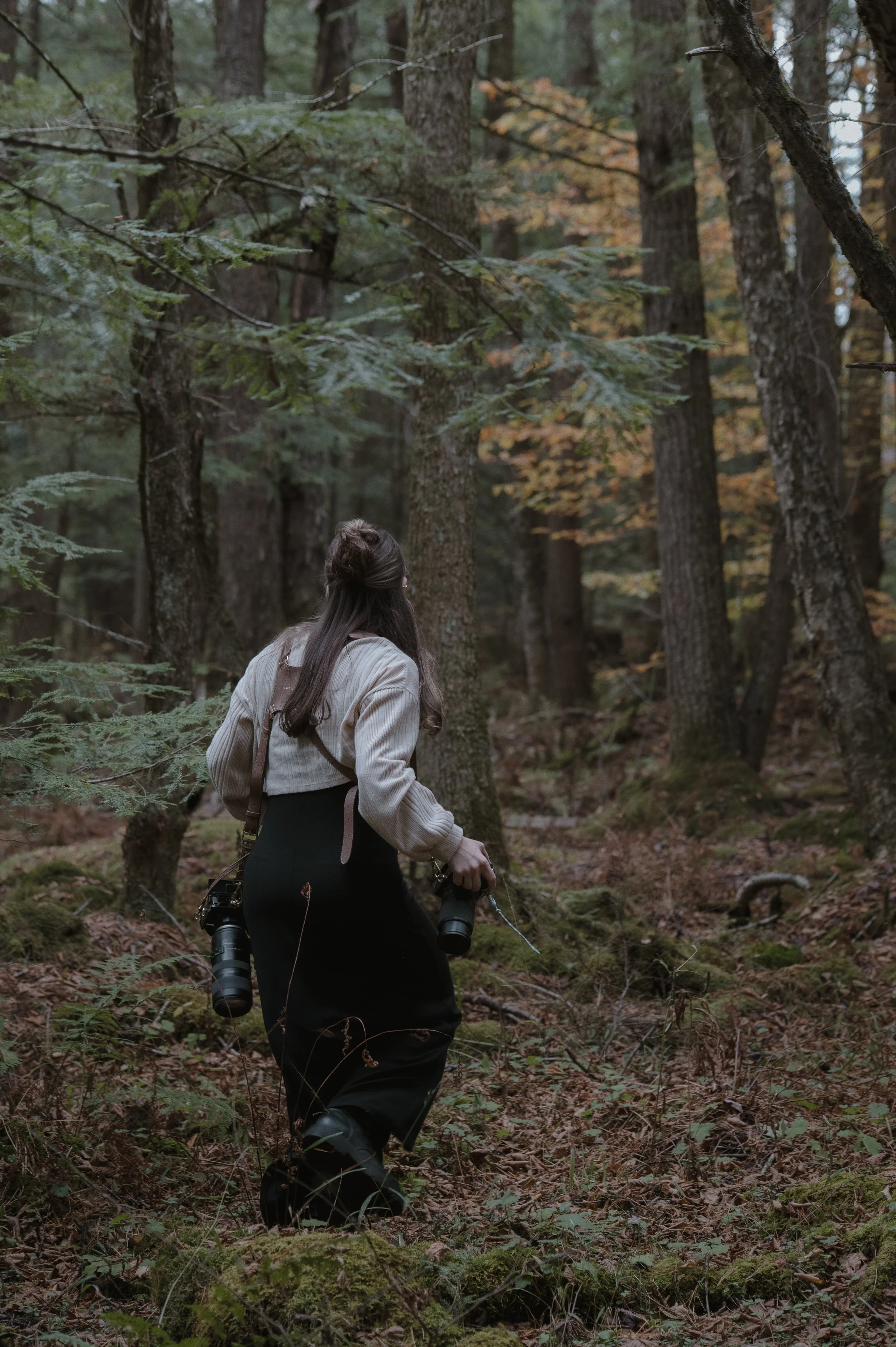 A woman walking through a forest with trees and fallen leaves, carrying a camera and binoculars.