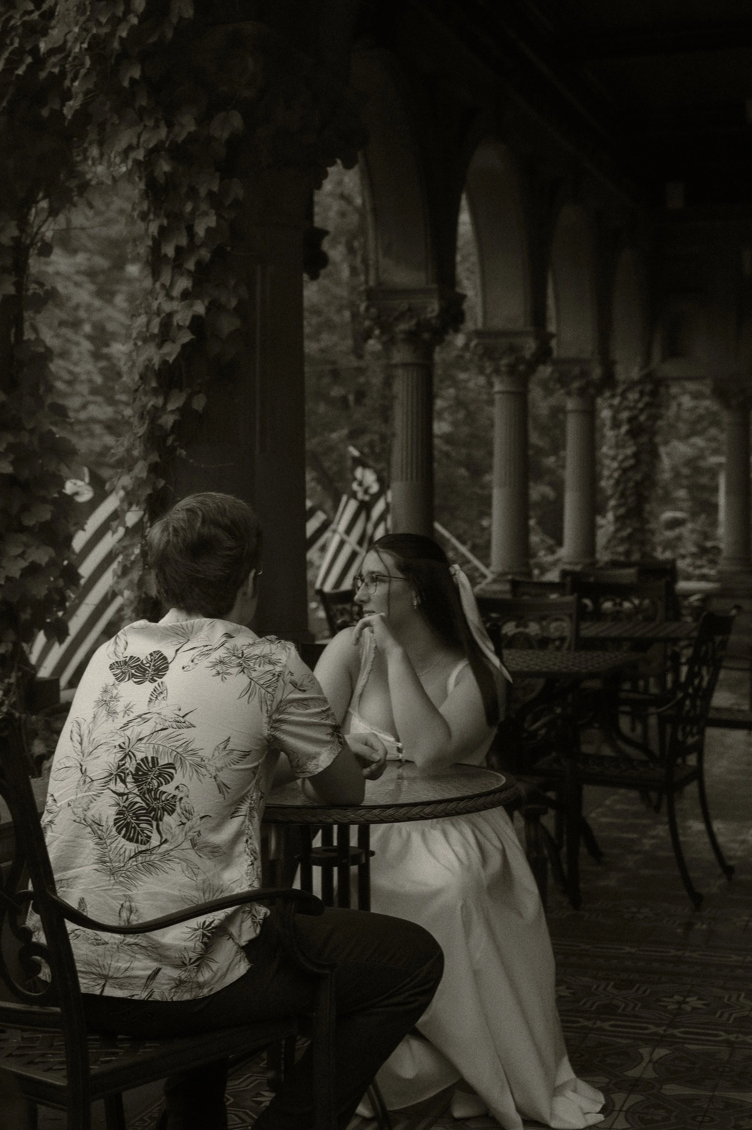 A man and woman sitting at a table in a covered outdoor setting, having a conversation. The woman is wearing a white dress and glasses, while the man is wearing a Hawaiian shirt. There are ivy-covered columns and American flags in the background.