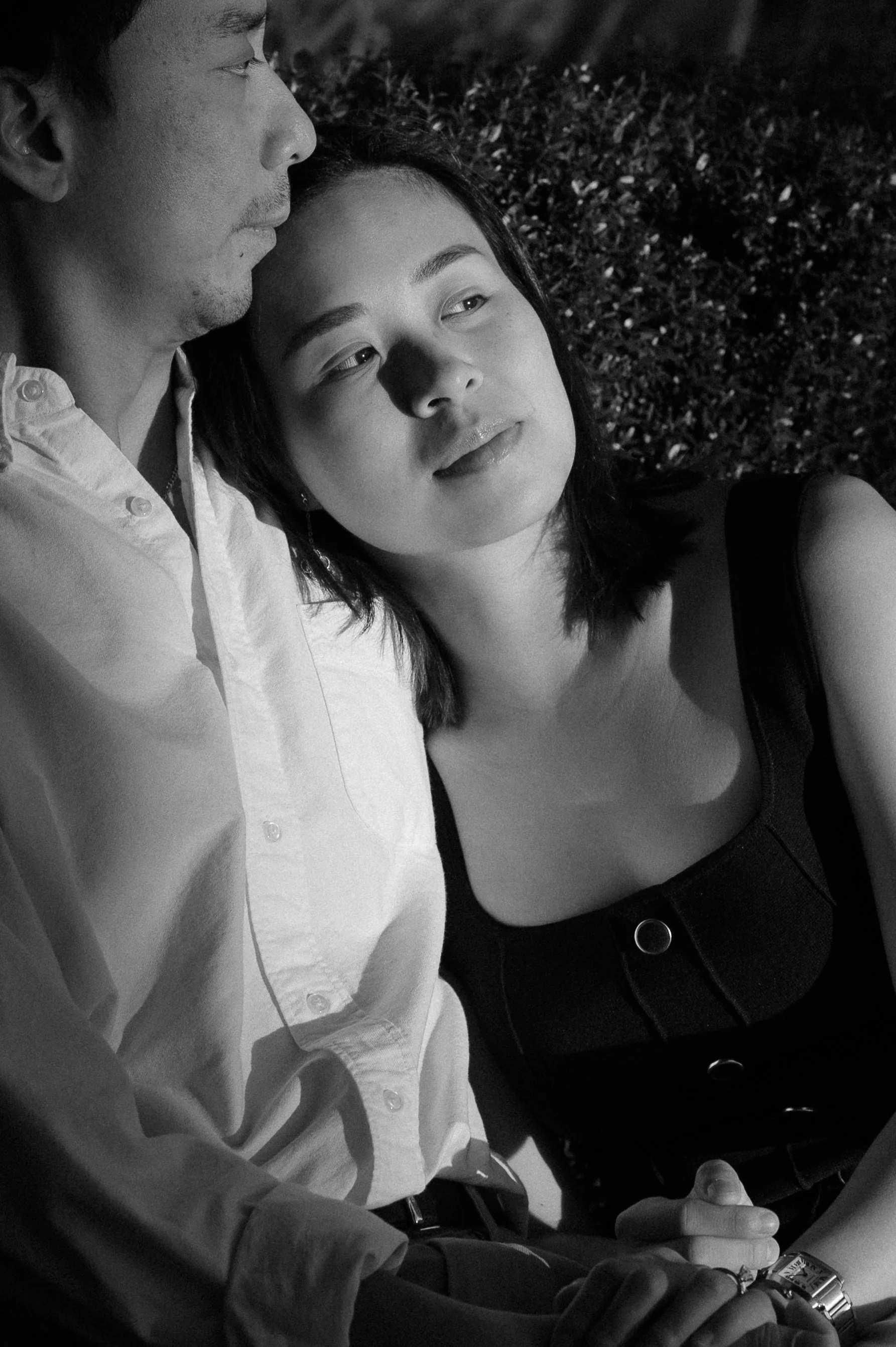 A black and white photo of a young woman resting her head on an older man’s shoulder, both sitting outdoors with bushes in the background. The woman looks slightly to the side with a peaceful expression, and the man’s face is partially visible.