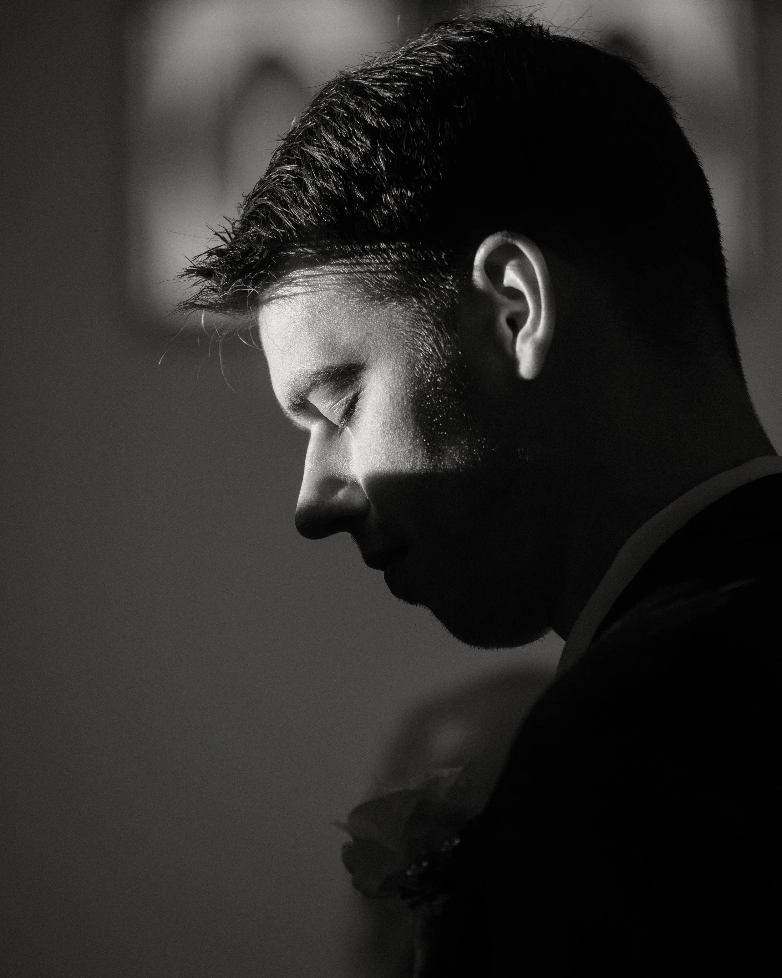 Black and white close-up side profile of a man with eyes closed, with short, dark hair, and a beard, dressed in formal attire, with soft lighting highlighting his facial features.