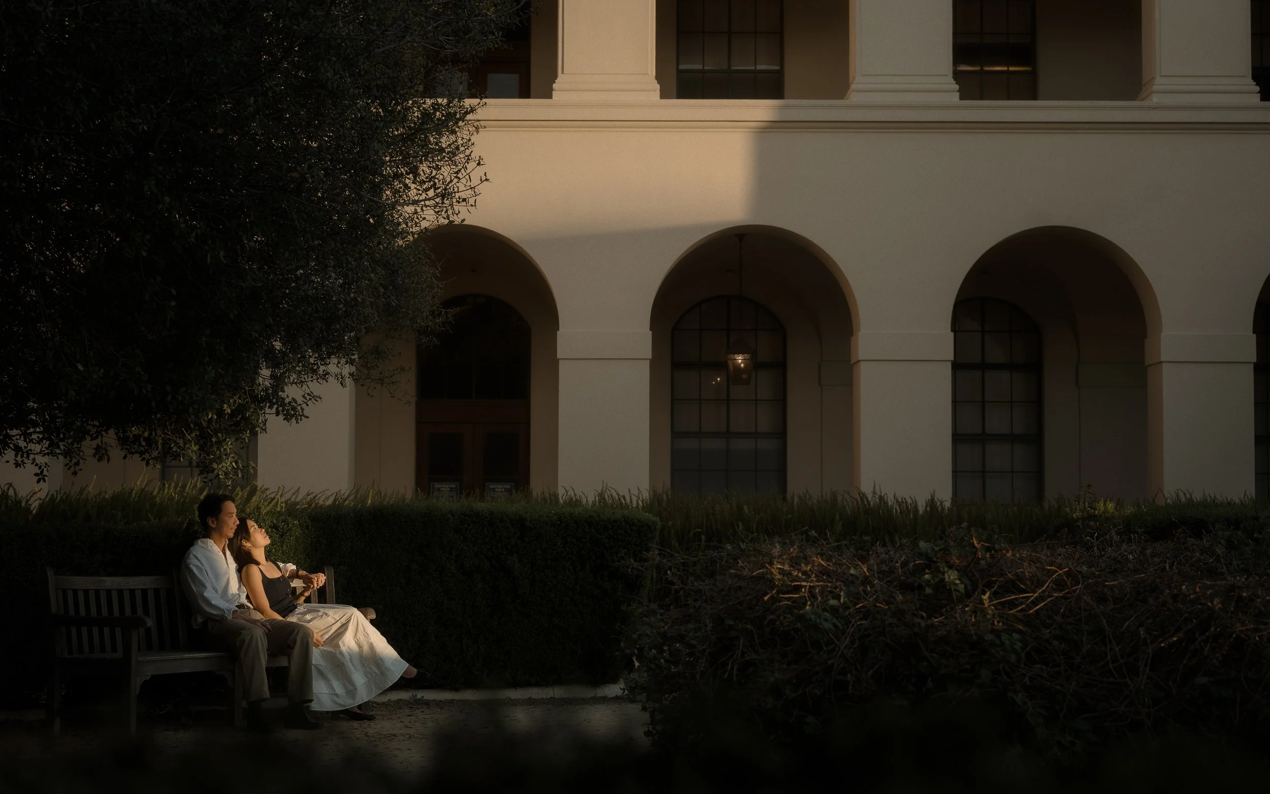 Two people sitting on a park bench in front of a large building during dusk. The woman is wearing a white dress and the man is dressed in a white shirt and dark pants. They are looking up, possibly at the sky or something in the distance.
