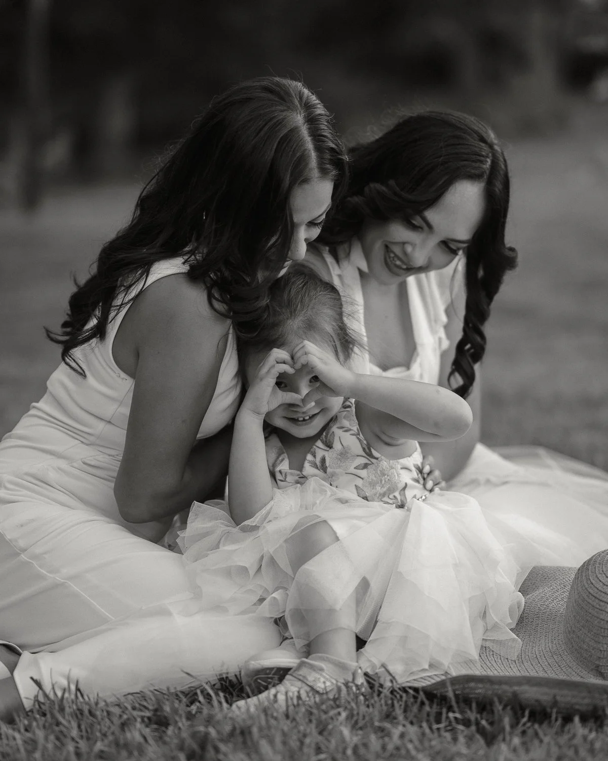 Two women and a young girl sitting outdoors on grass, smiling and making playful gestures, in black and white.