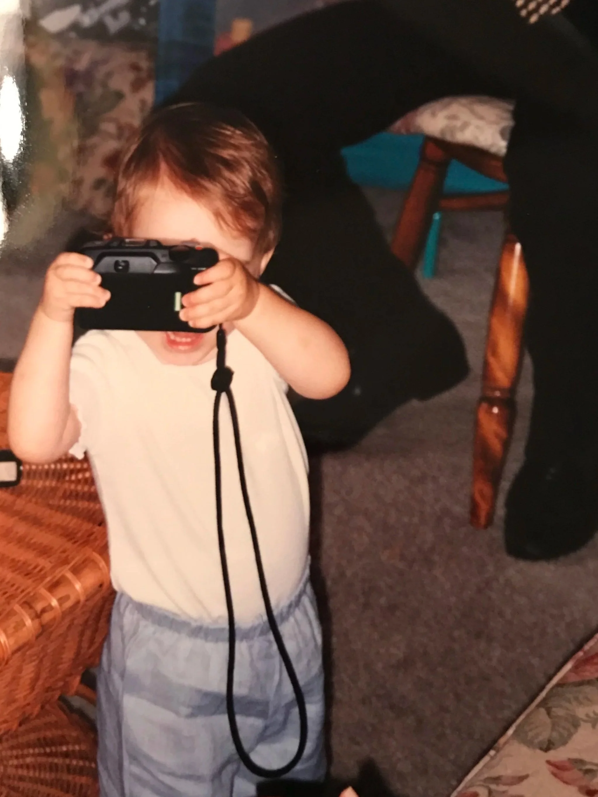 A young child with brown hair taking a photo with a camera, standing indoors near a wicker chair, with an adult person partially visible behind him and furniture in the background.
