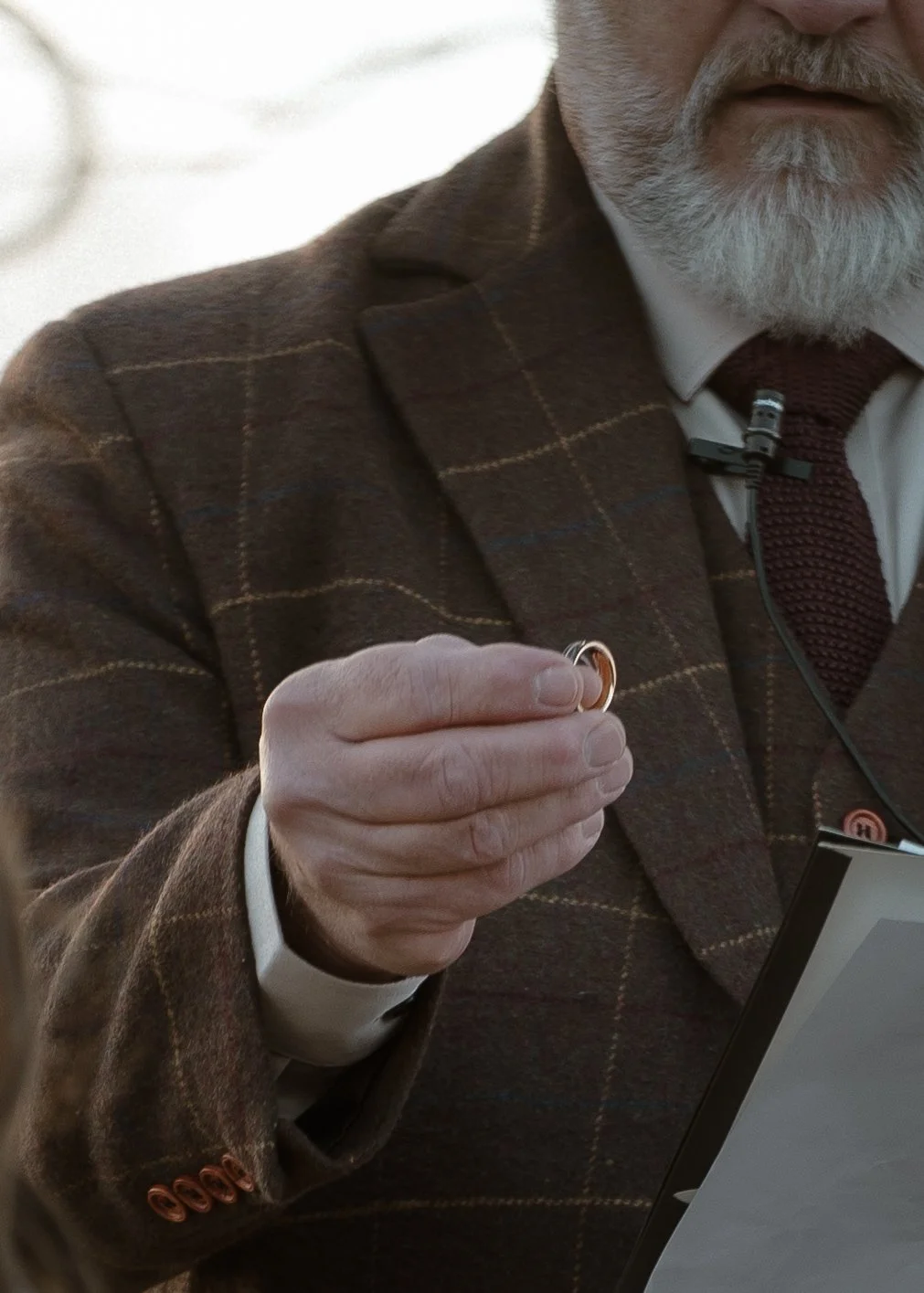 A man with a gray beard wearing a checkered brown blazer, white shirt, and maroon tie, holding a small gold ring in his hand. He is also wearing a microphone and has a computer or tablet in front of him.