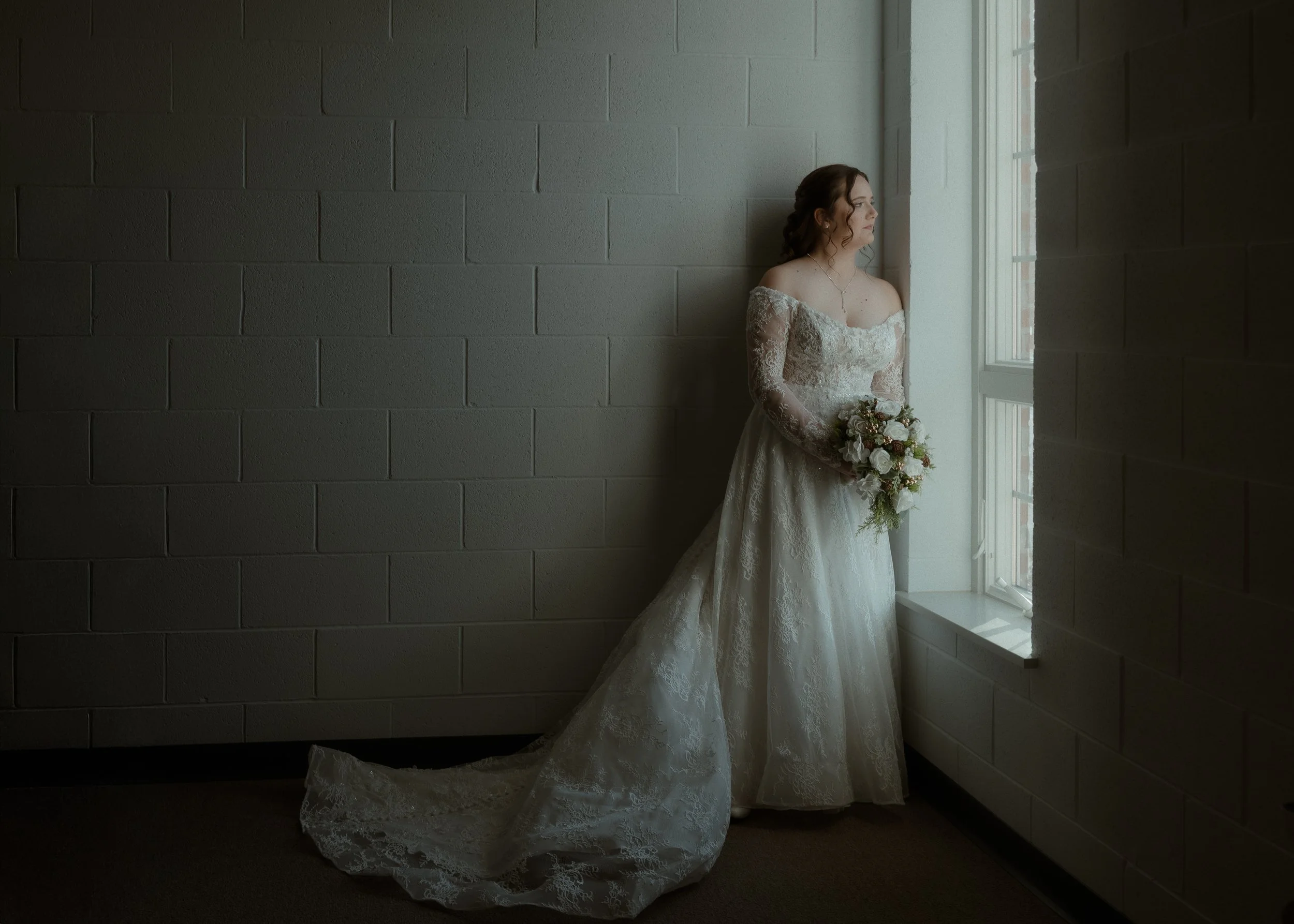 A bride in a lace wedding dress holding a bouquet, standing by a window with soft natural light illuminating her face.