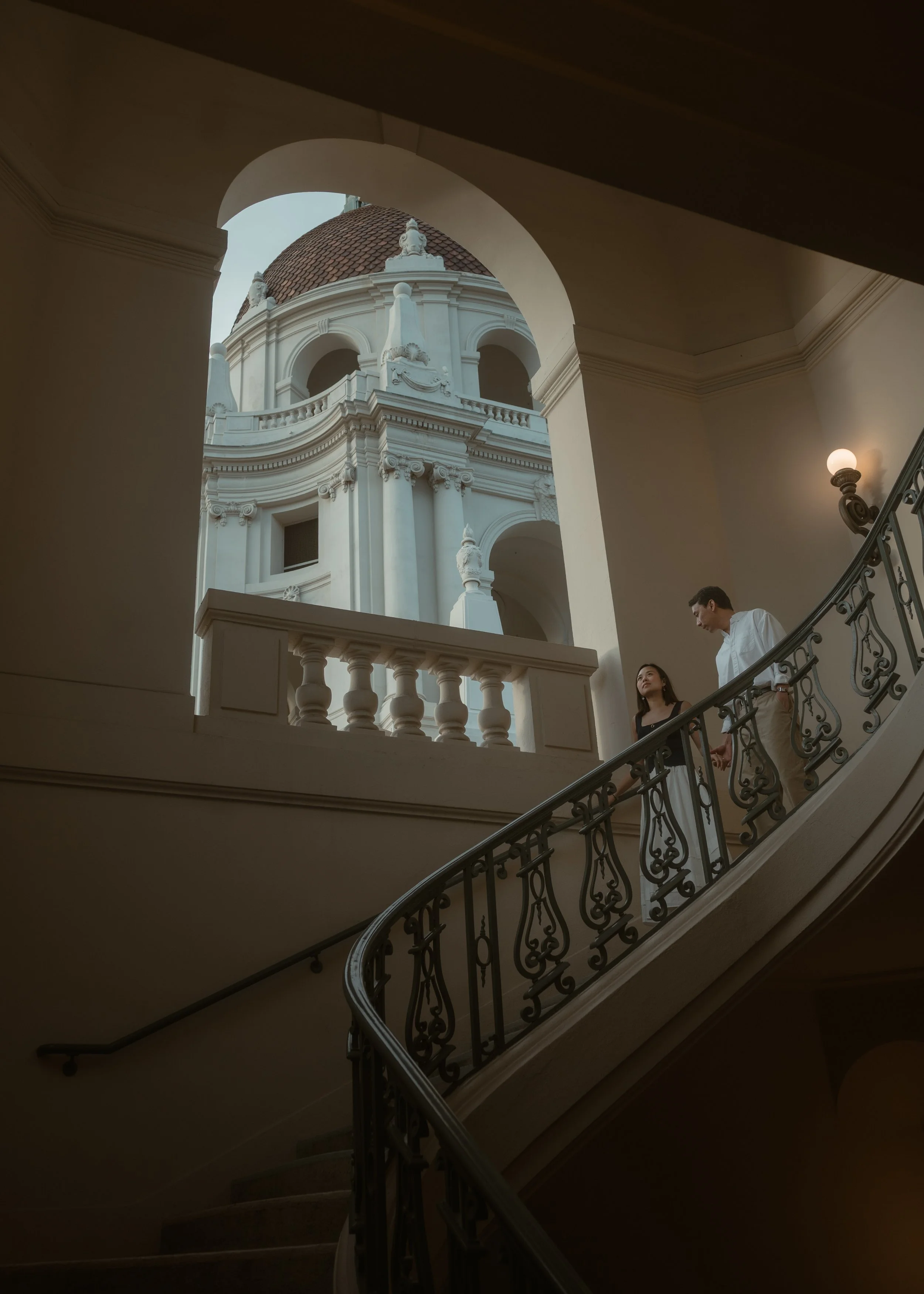 A man and woman walking down an ornate staircase inside a building with classical architecture, looking at each other. Through an archway, a large domed building with detailed white architecture is visible outside.