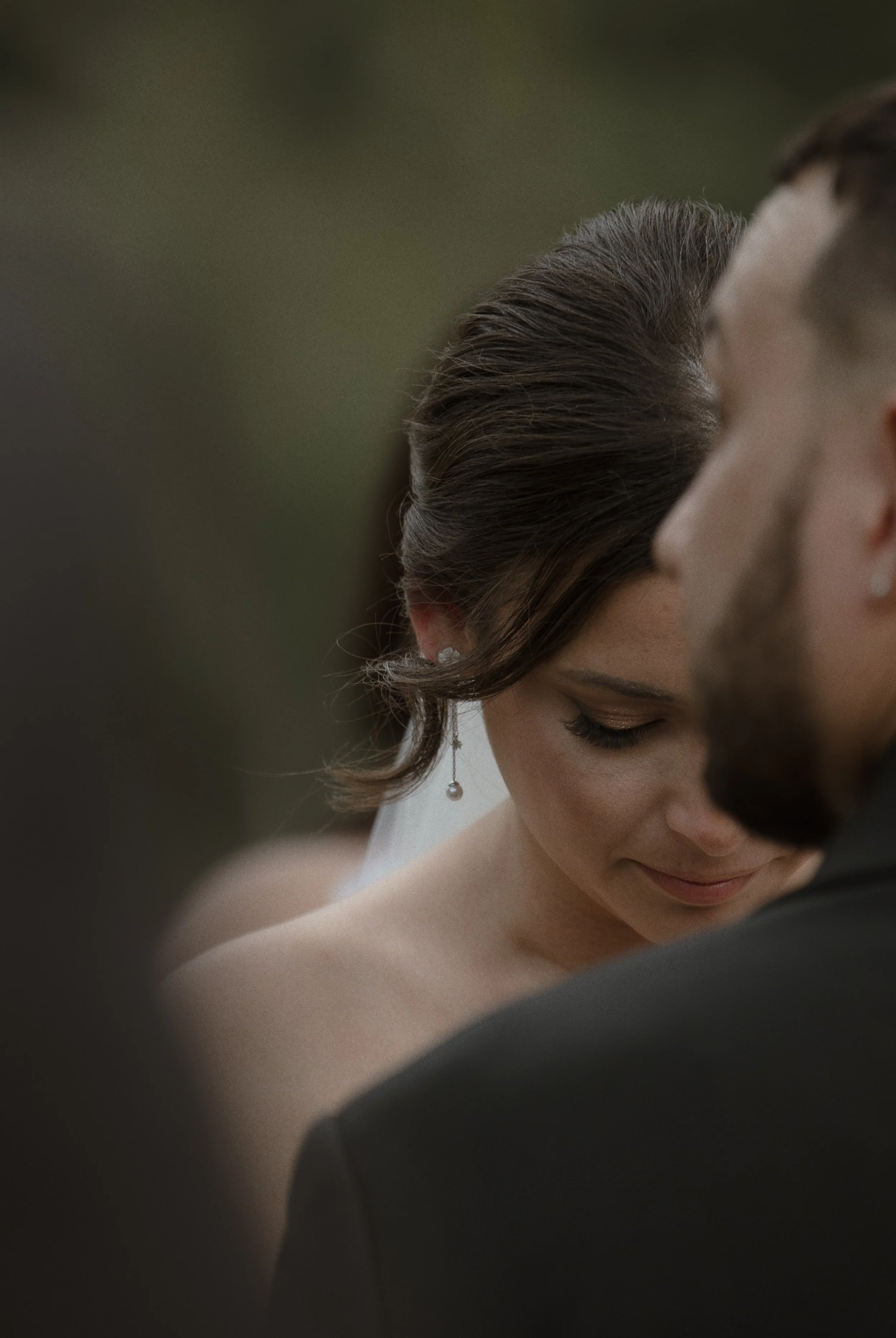 Close-up of a woman with dark hair styled in an updo, wearing earrings and a veil, smiling with her eyes closed, standing close to a man during a wedding ceremony.