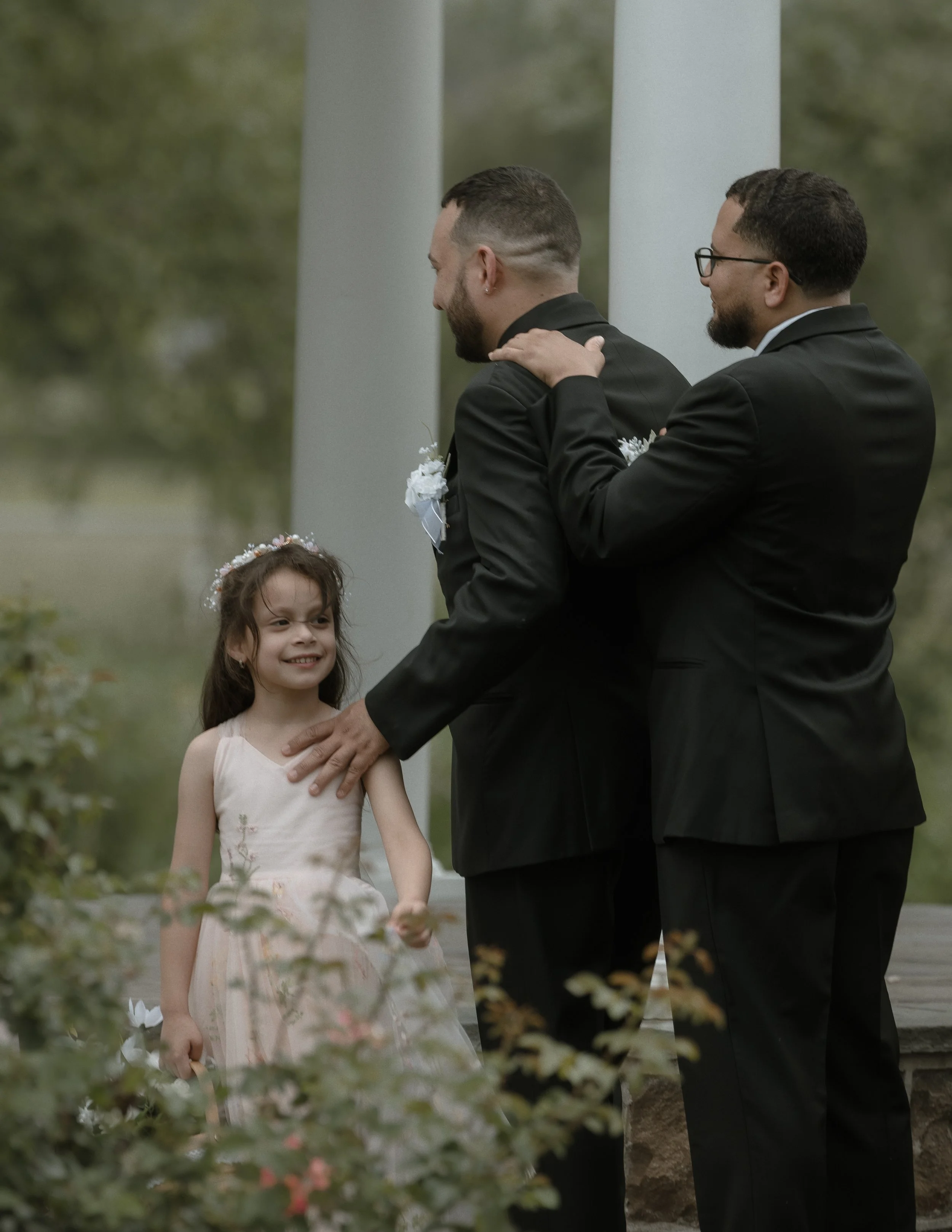 Two men in black suits, one with glasses, and a young girl in a light pink dress, outdoors near a white pavilion, with greenery in the background. The girl has a flower crown, and the men appear to be participating in a formal or ceremonial event.